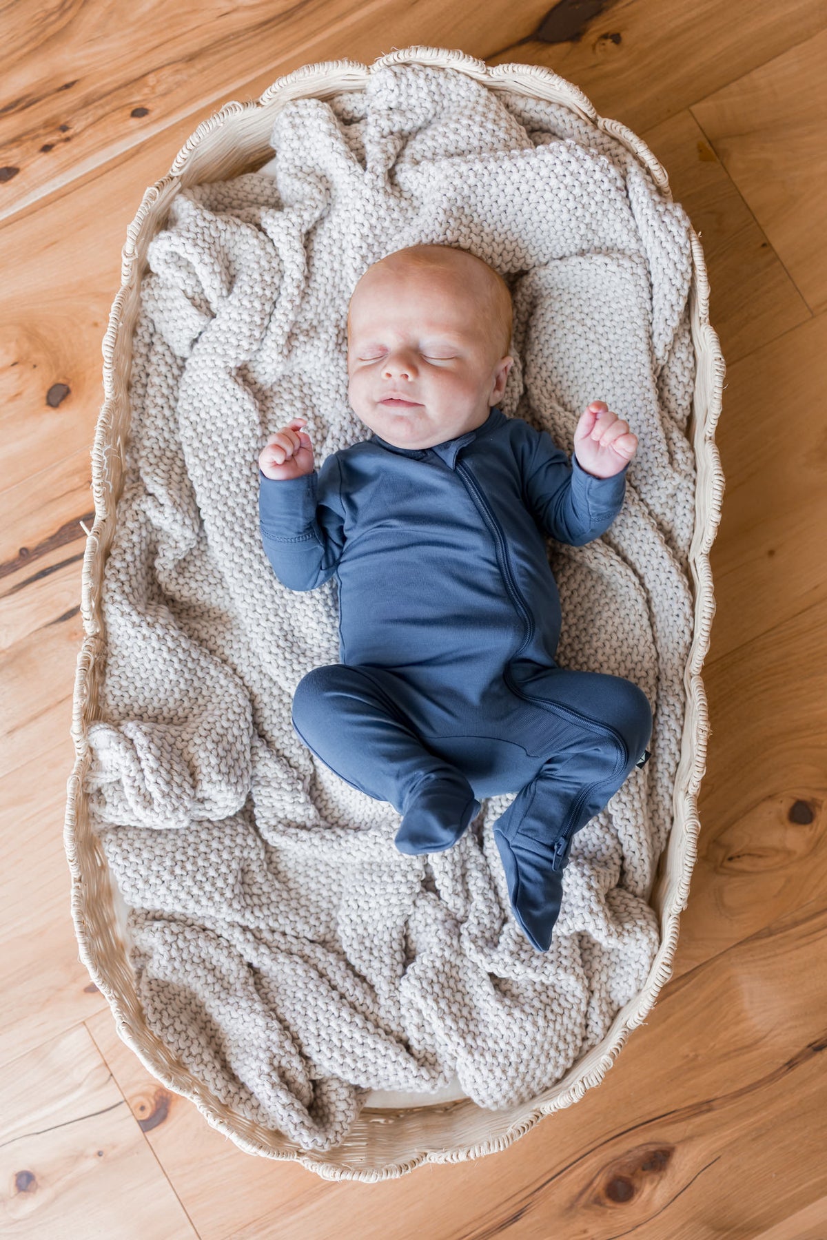 A sleeping newborn baby wearing a navy blue footed zipper onesie, lying on a textured cream knit blanket inside a woven oval bassinet on a wooden floor.