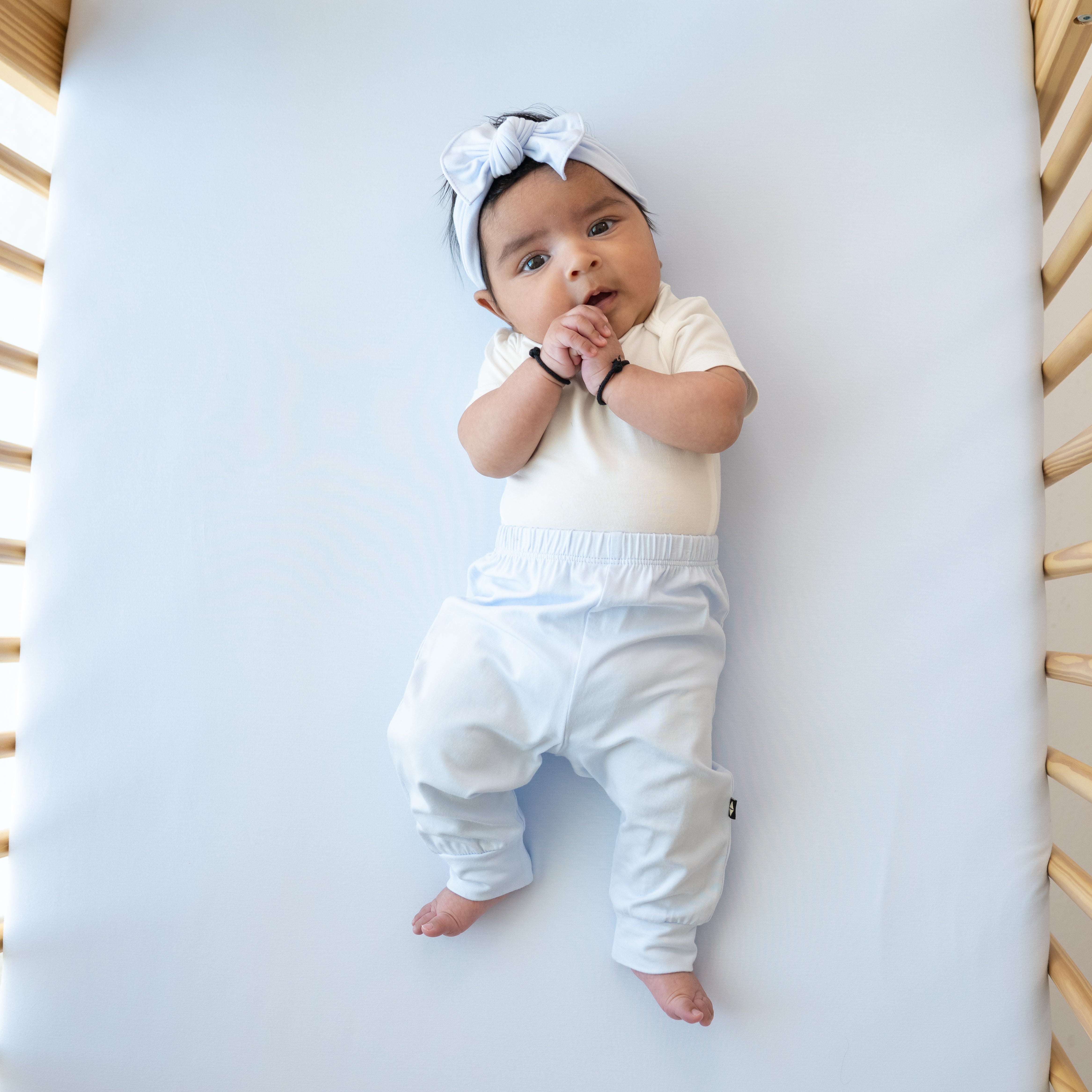 Infant girl laying on a Mist Crib Sheet wearing a Knotted Bow Headband in Mist with matching Mist pants and a Cloud Bodysuit