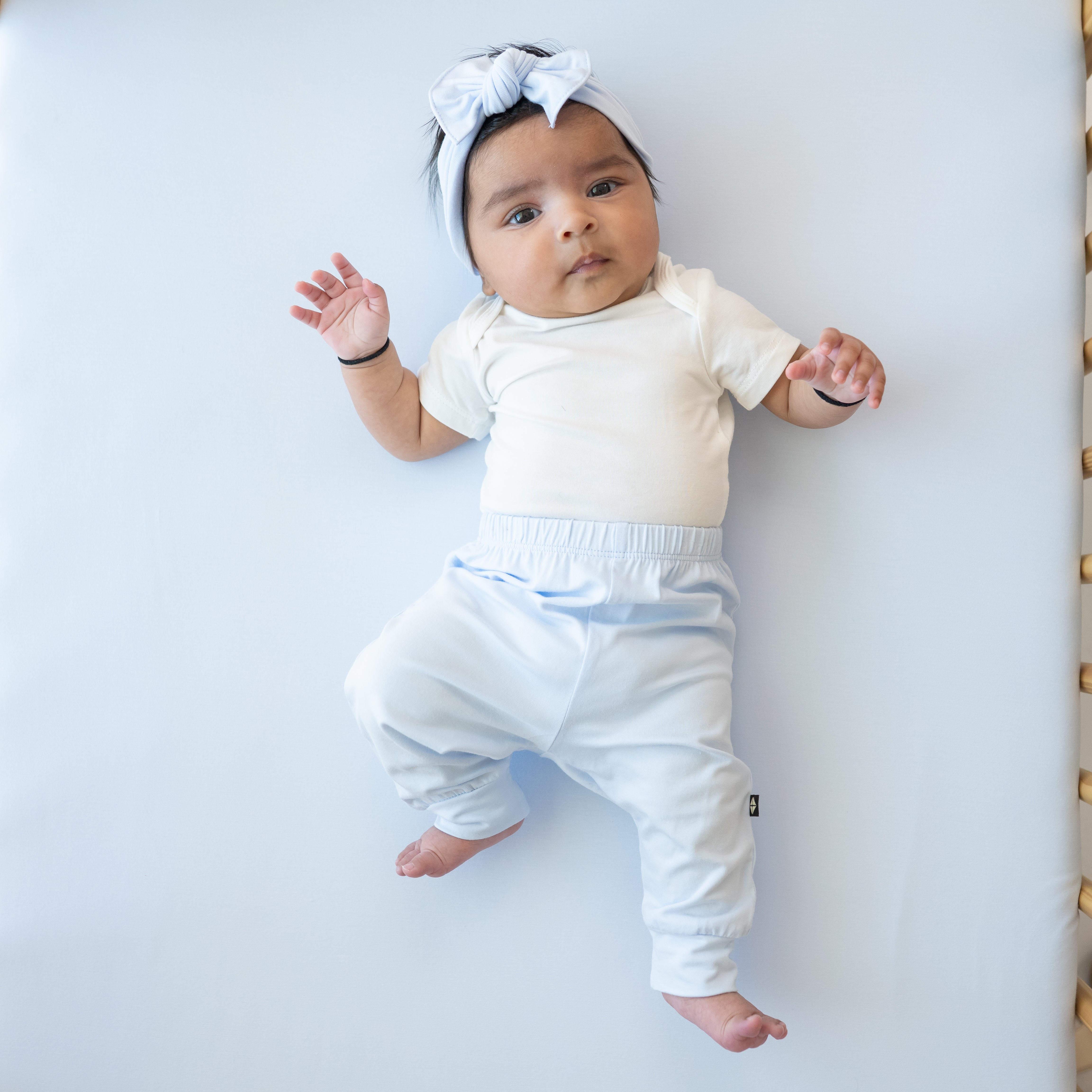 Infant model laying in a crib wearing the Pant in Mist and matching knotted bow with a Cloud Bodysuit