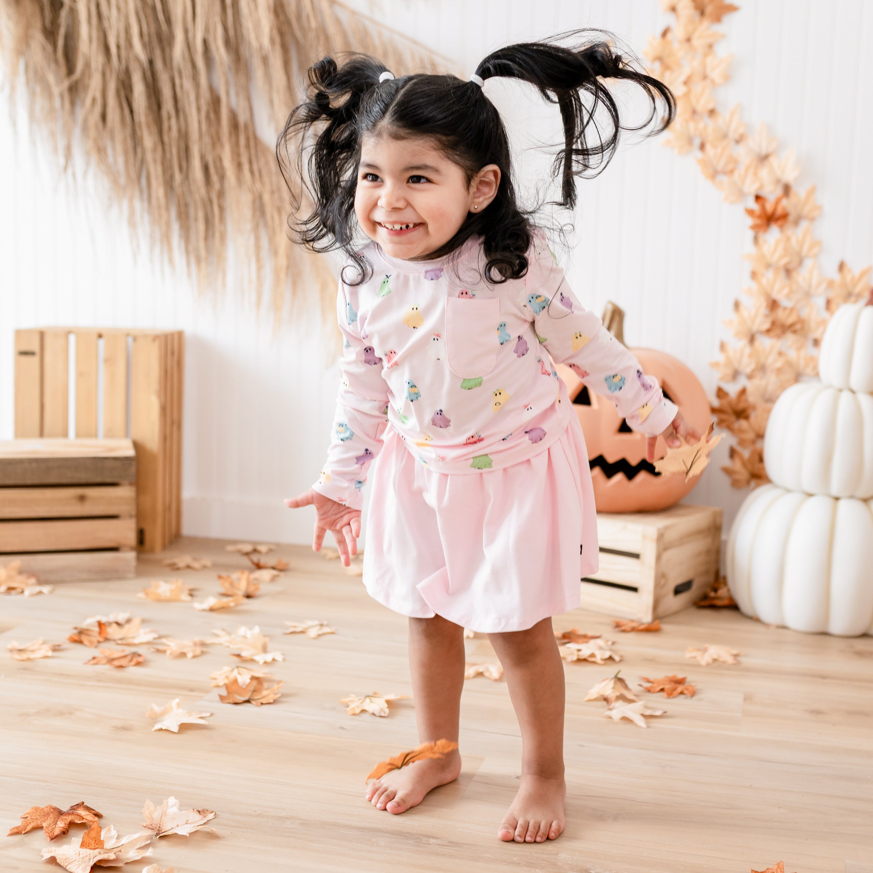 Smiling toddler wearing the Long Sleeve Toddler Crew Neck Tee in Pastel Ghost paired with the Sakura skort in front of a fall backdrop of leaves, pumpkins and wooden crates