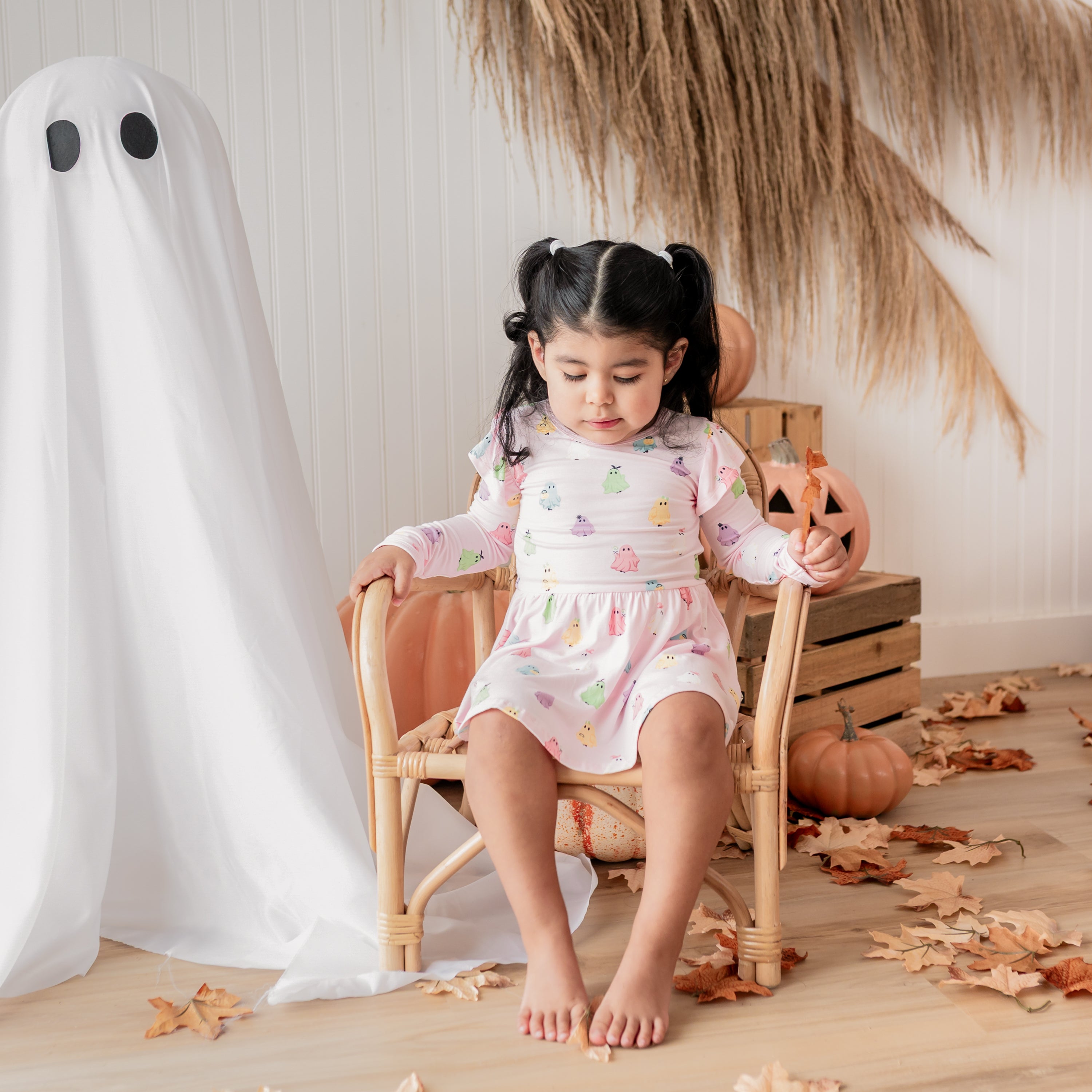 Toddler girl sitting in a wicker chair beside a sheet ghost and in front of a fall backdrop of wheat, pumpkins, leaves and wooden crates wearing the Long Sleeve Twirl Bodysuit Dress in Pastel Ghost
