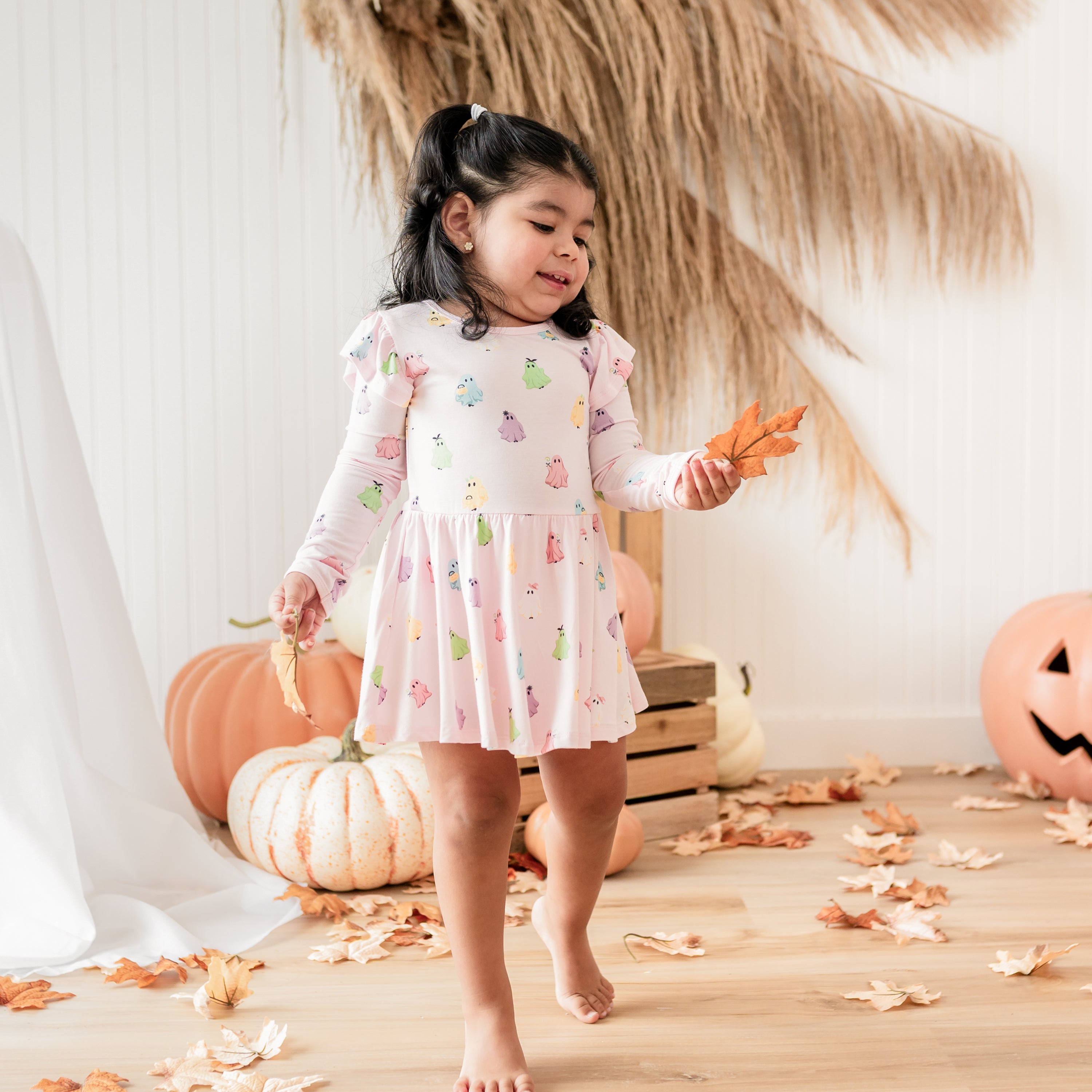 Toddler standing holding leaves in her hand walking wearing the Long Sleeve Twirl Bodysuit Dress in Pastel Ghost in front of a fall back drop of pumpkins, leaves and a wooden crate