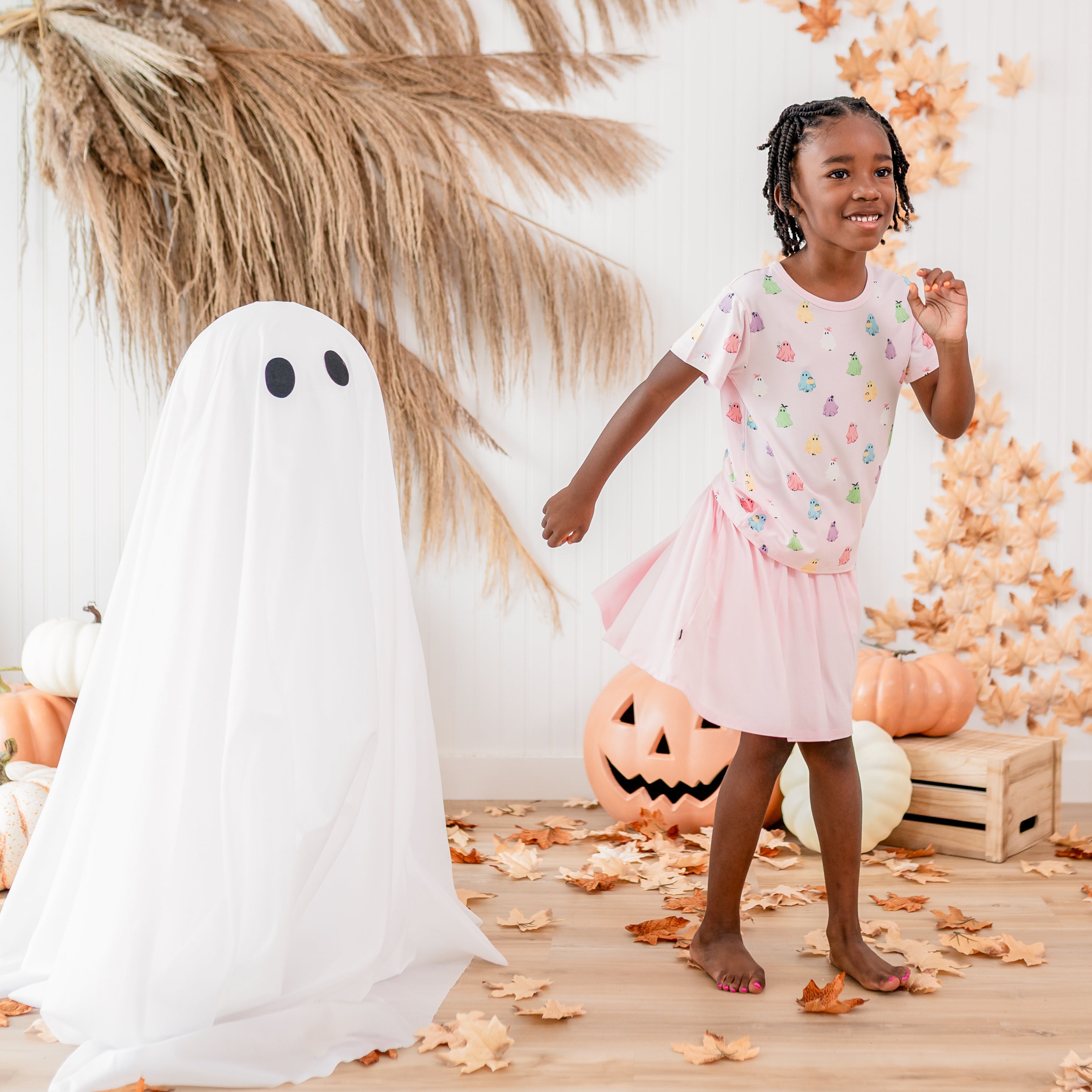 Smiling girl twirling in the Toddler Basic Tee in Pastel Ghost and Sakura Skort standing beside a sheet ghost and in front of a fall backdrop