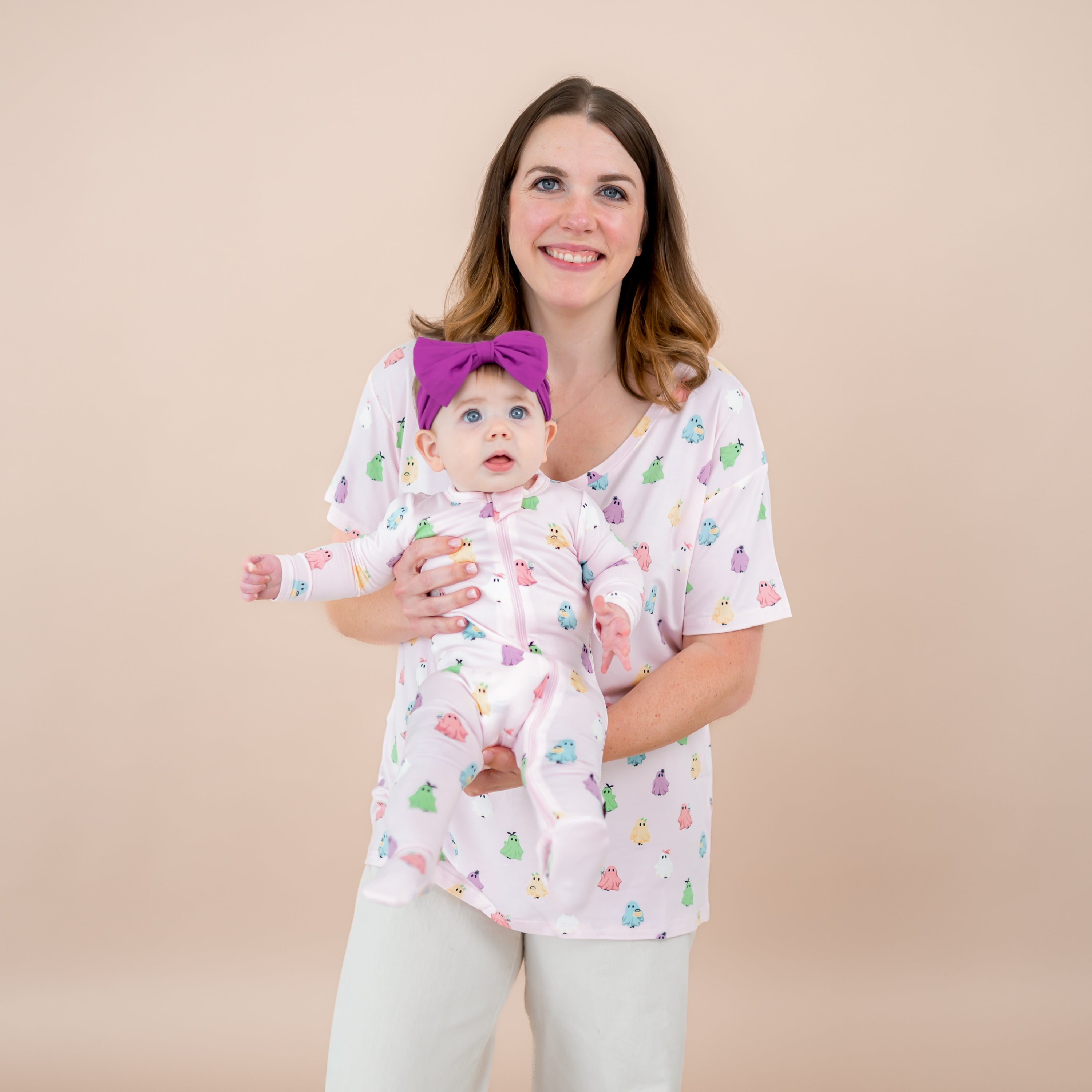Mother and daughter matching in Pastel Ghosts from the Halloween collection. Mother wearing the Women's Relaxed Fit V-Neck in Pastel Ghost holding her infant daughter in matching zippered footie paired with a Berry Bow Headband