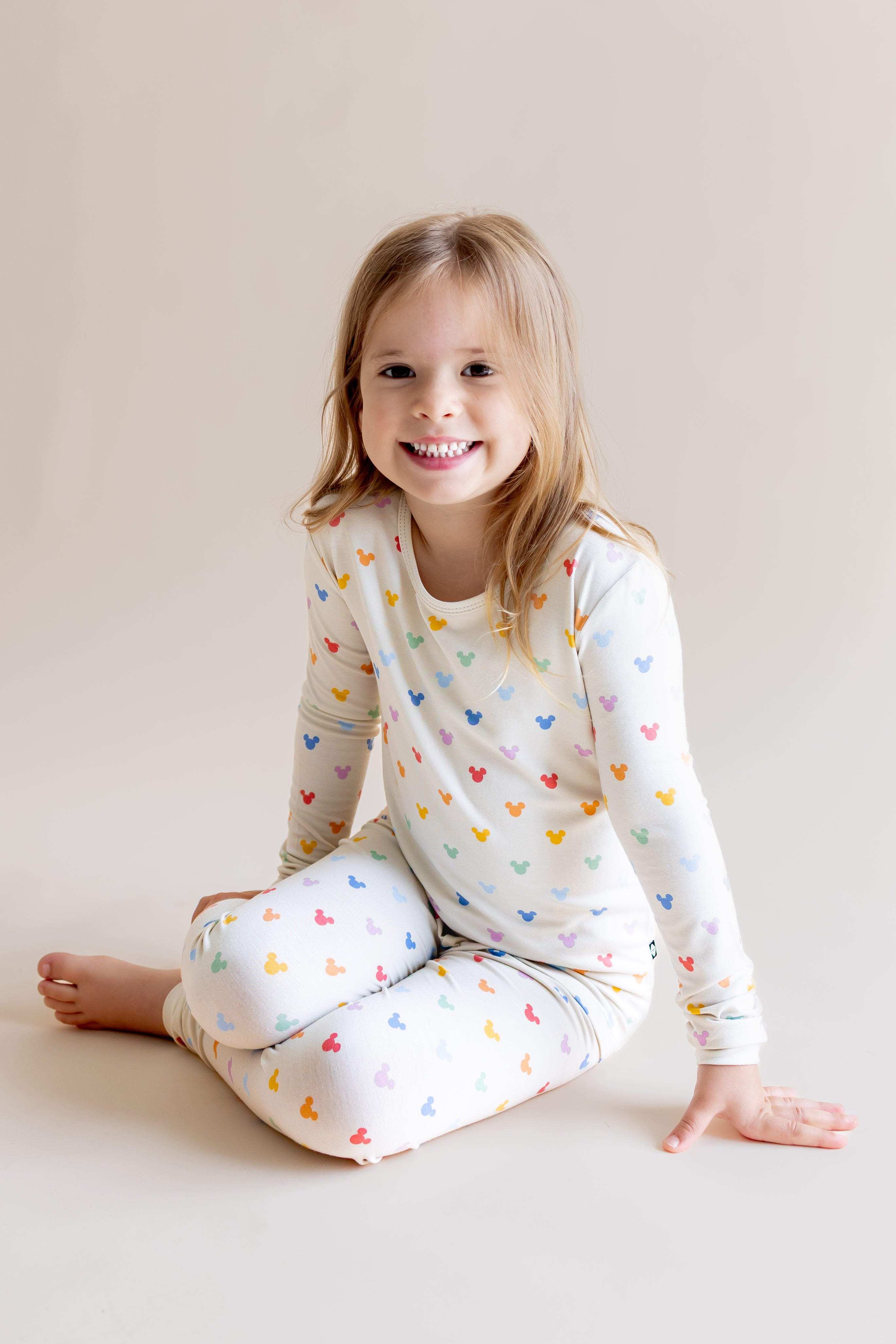 Girl sitting in Long Sleeve Pajamas in Pastel Mickey Mouse print on a cream background