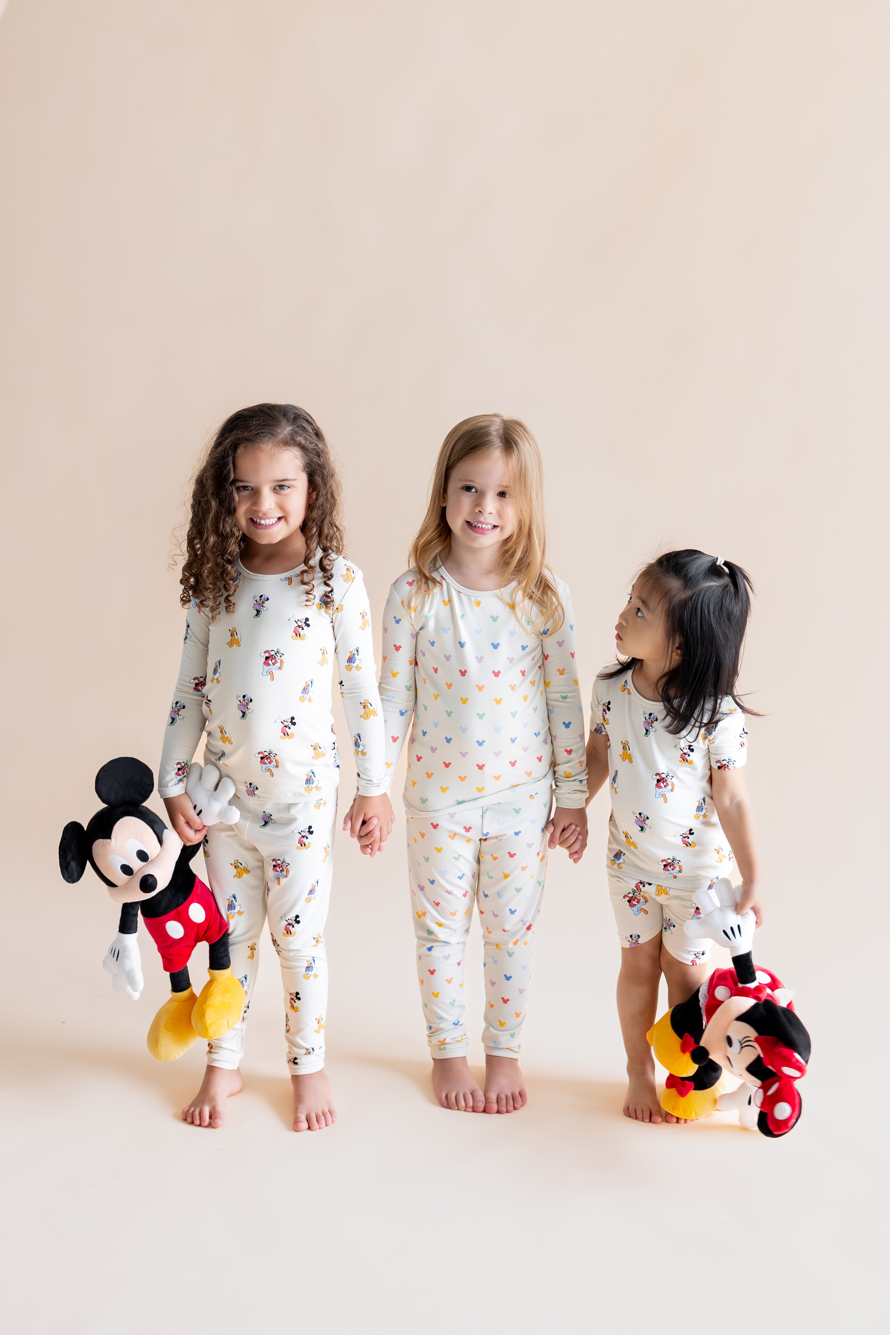 3 Girls standing holding each others hands wearing Long Sleeve Pajamas in in Mickey Mouse & Friends and Pastel Mickey Mouse print and Short Sleeve Pajamas in Mickey Mouse & Friends print holding stuff toys on a white background