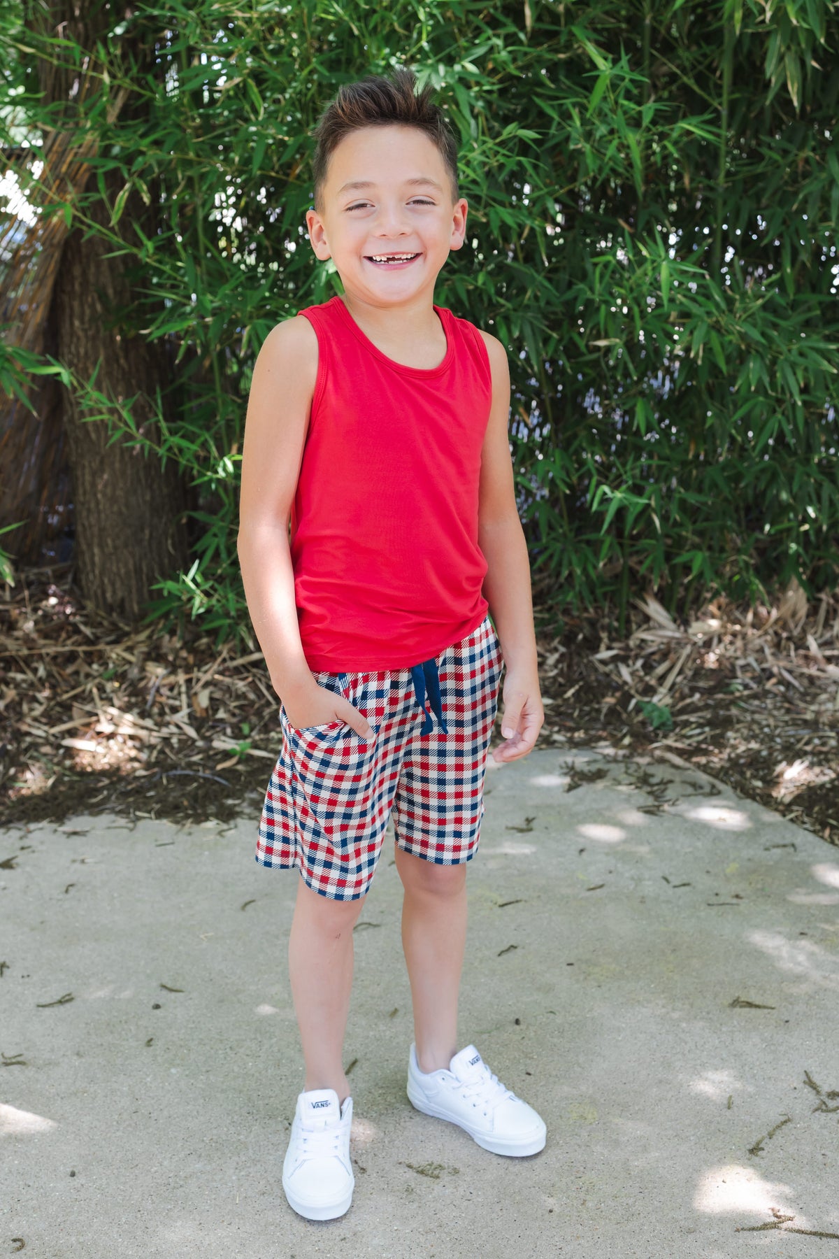 Child wearing a red blue and white Patriotic Gingham check print drawstring shorts outdoors over a red tank top and white shoes
