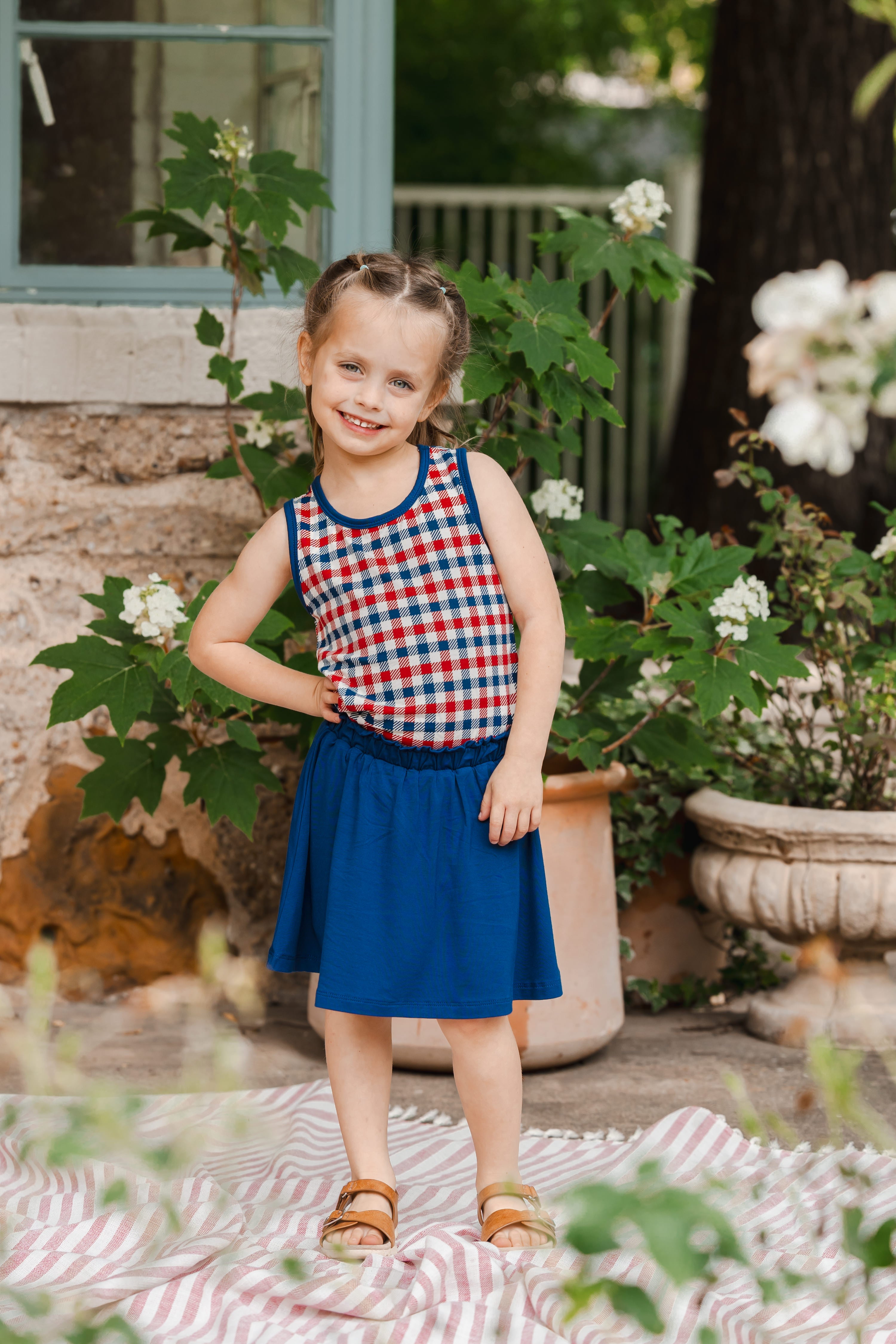 Child standing outdoors wearing a red blue and whtie Patriotic Gingham check print toddler tank top over a Tahoe skort and sandles