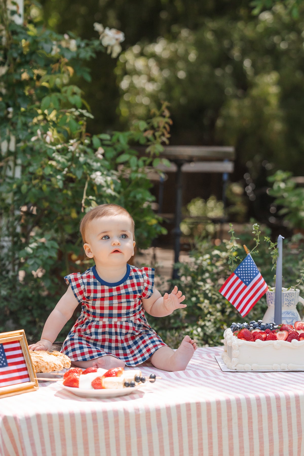 Child sitting outdoors on a table wearing a red blue and white Patriotic Gingham check print Twirl bodysuit dress