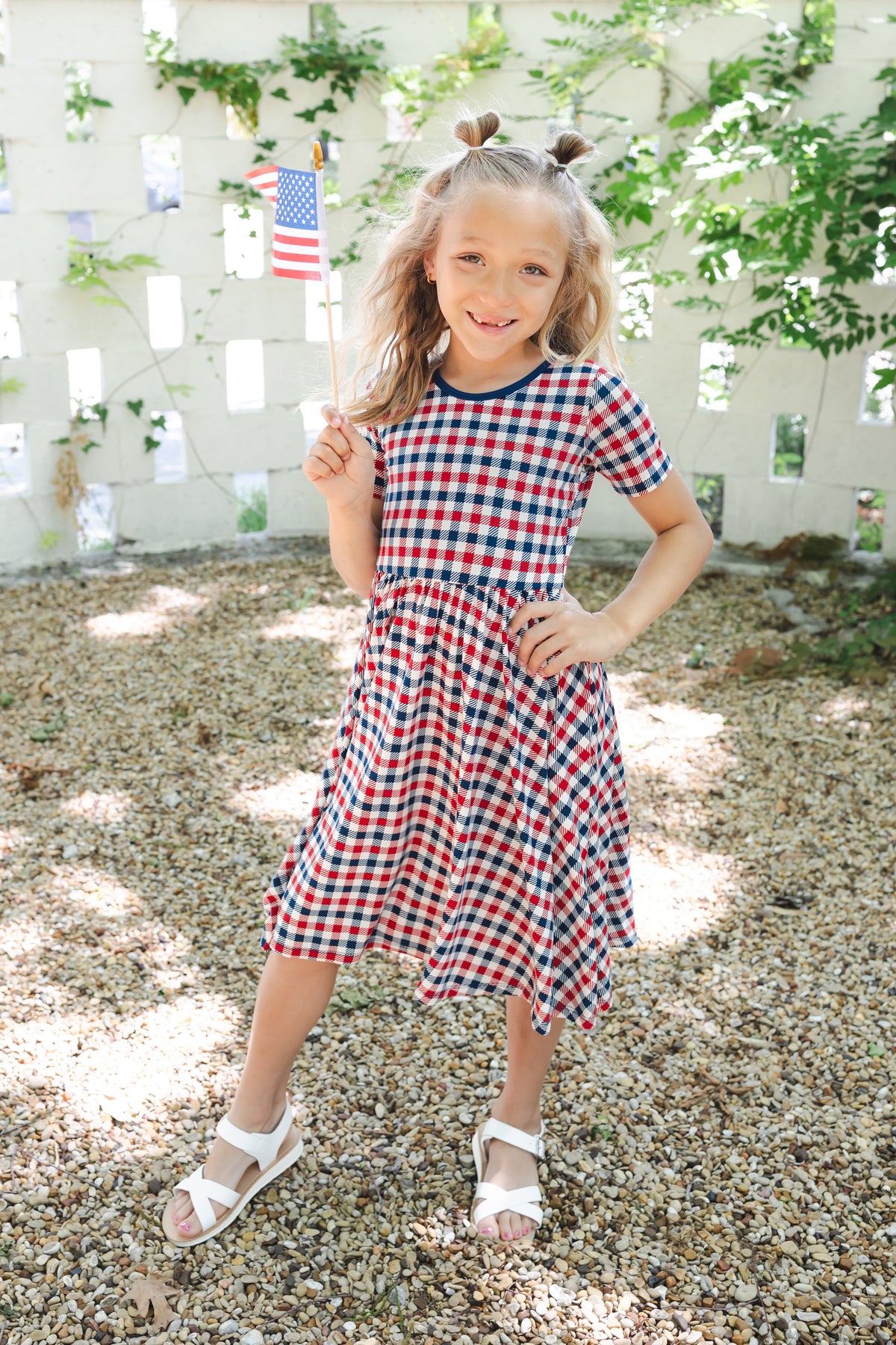 Child standing outdoors wearing a Red blue and white Patriotic Gingham check print Twirl Dress with white sandles holding a flag