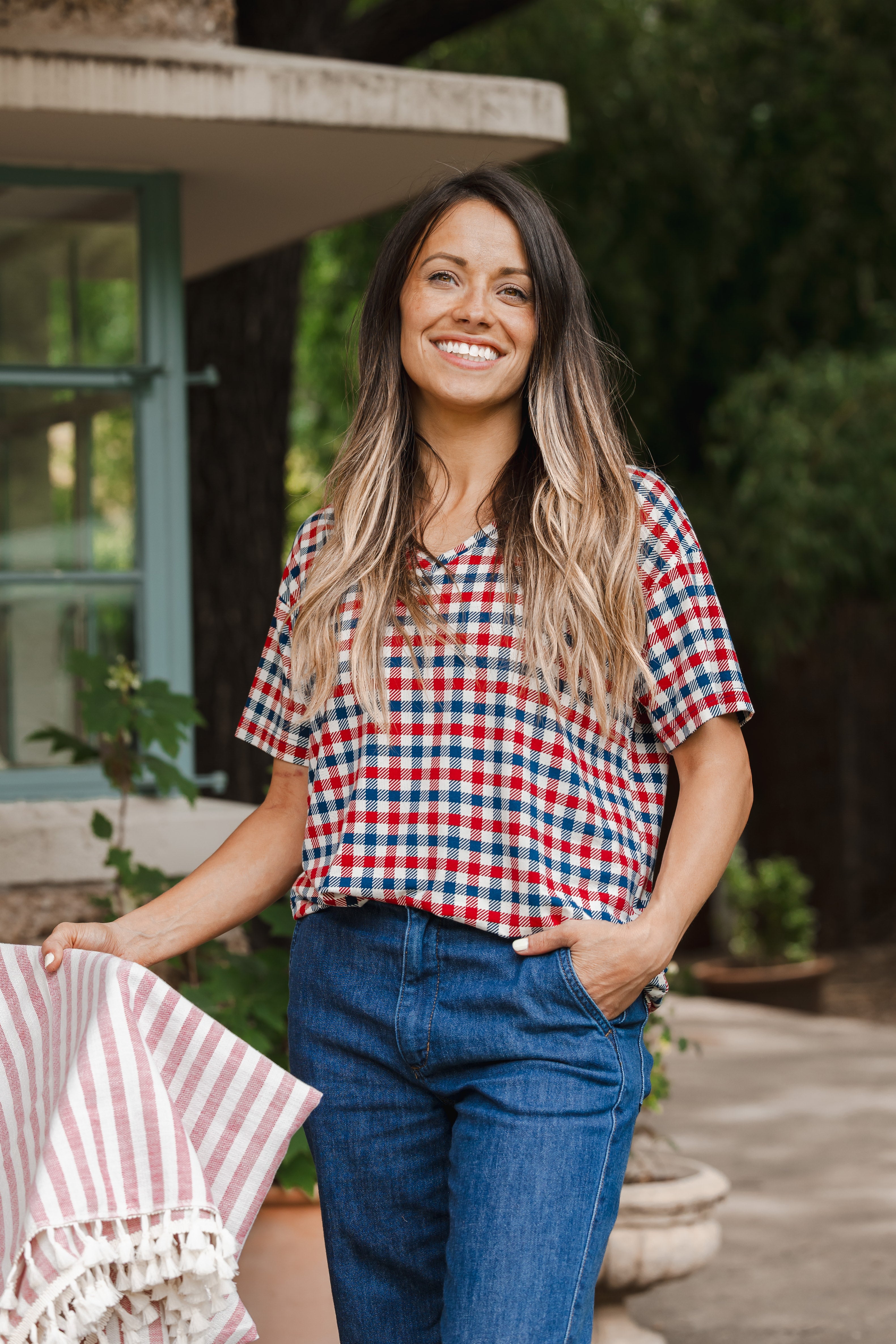Model standing outdoors wearing Women's Relaxed Fit V-Neck in red blue and white Patriotic Gingham check print over denim pants