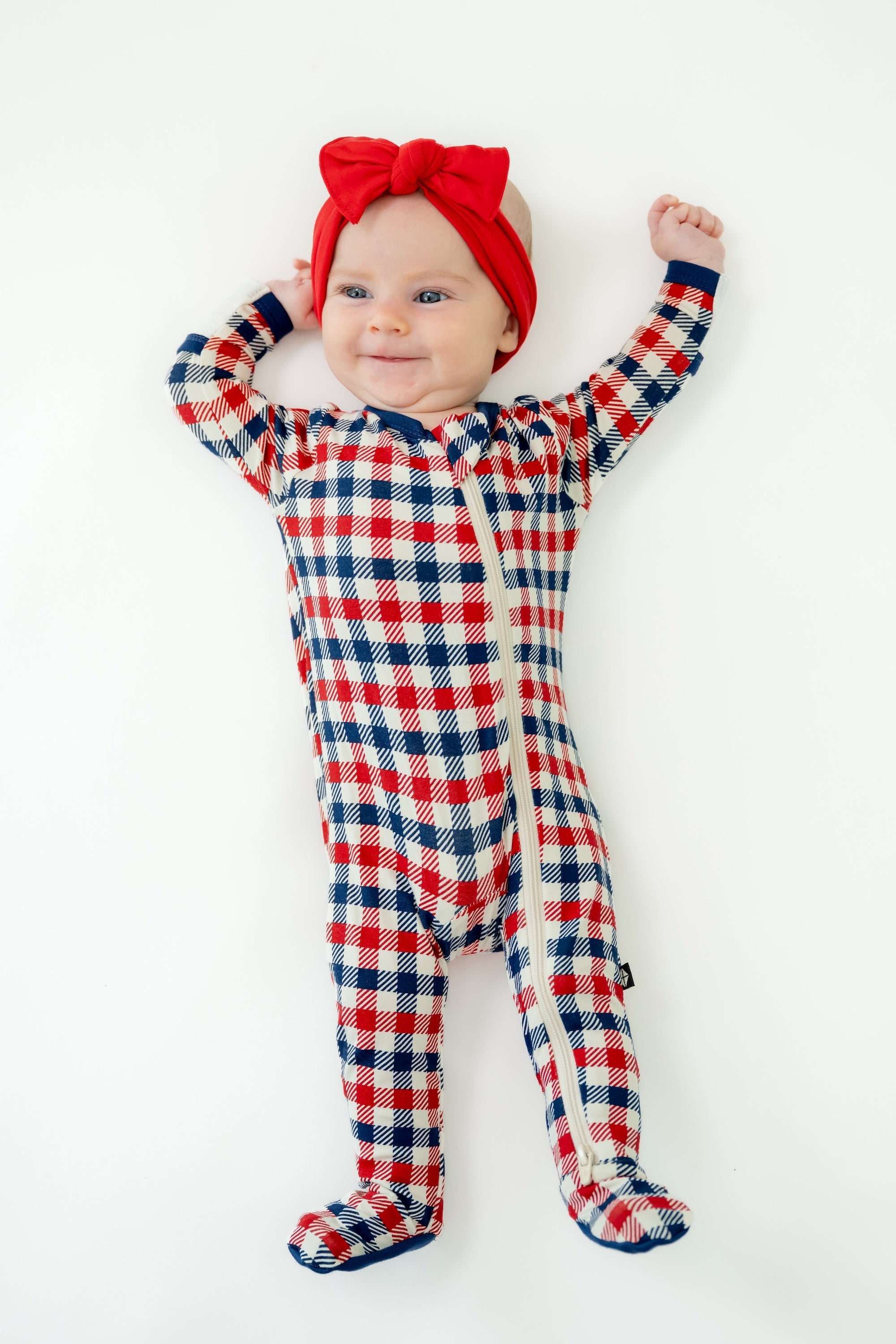 Baby wearing red white and blue Patriotic Gingham check print zippered footie with a red bow headband on a white background
