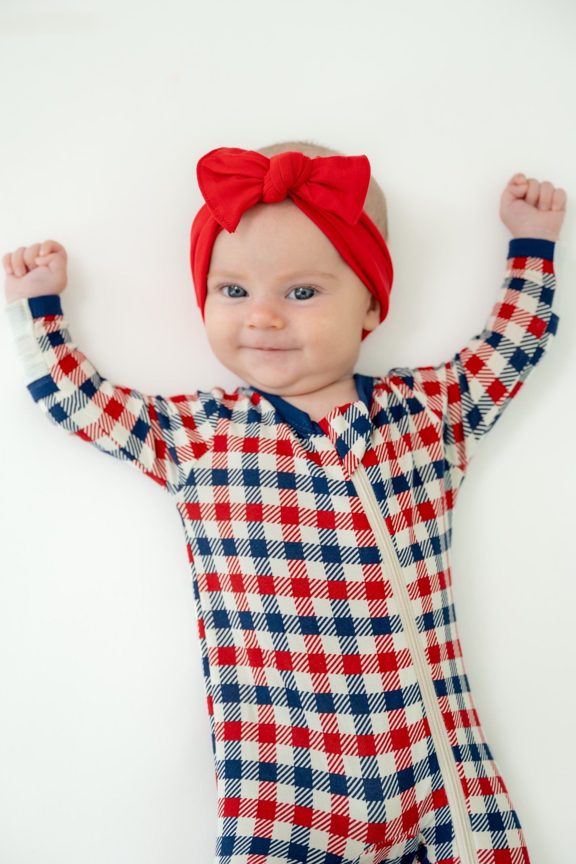 Baby in red white and blue Patriotic Gingham check print zippered footie with a red bow headband on a white background