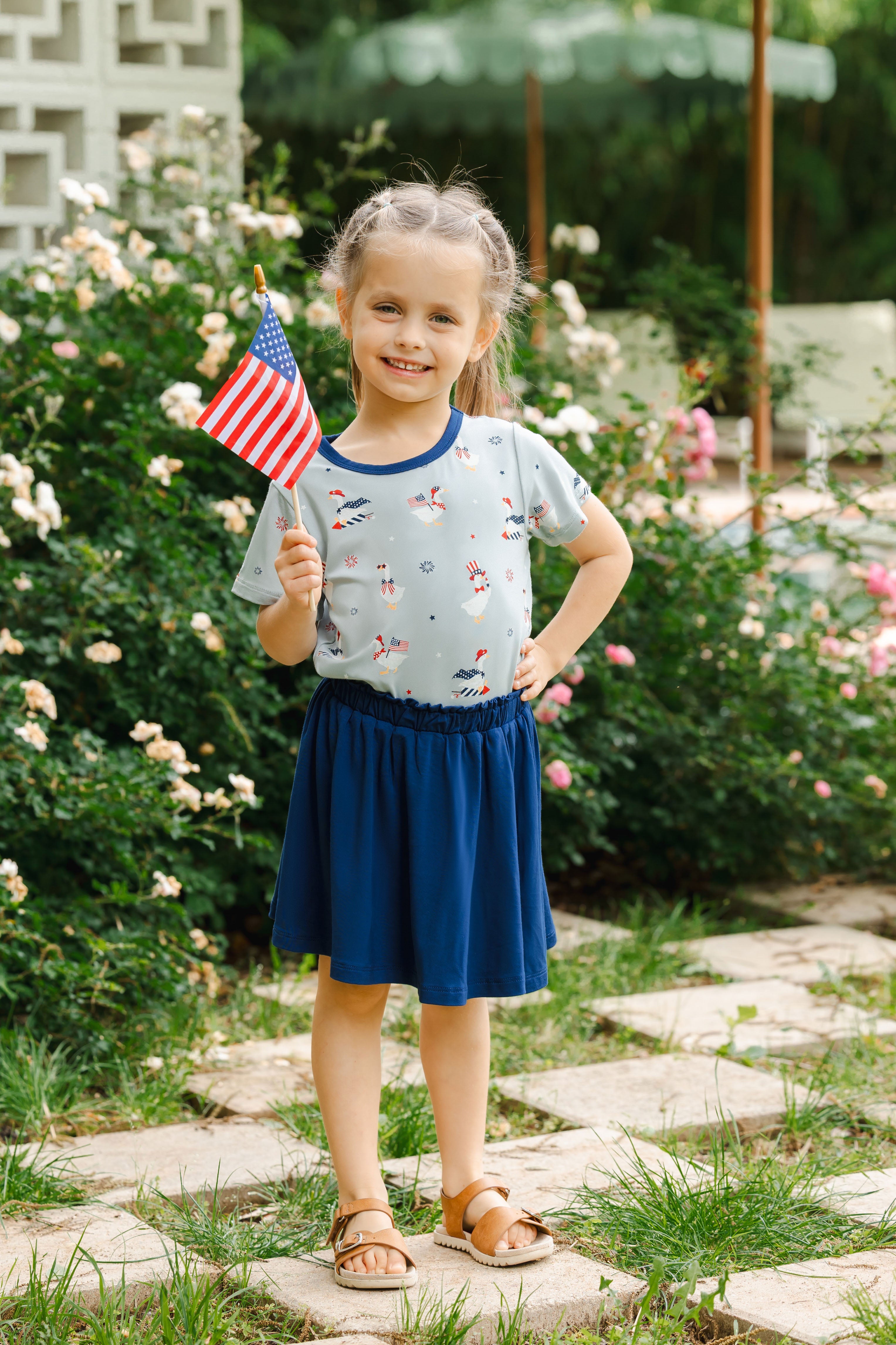 Child wearing light blue Toddler Basic Tee in Patriotic Goose print over a Tahoe skort holding a flag outdoors