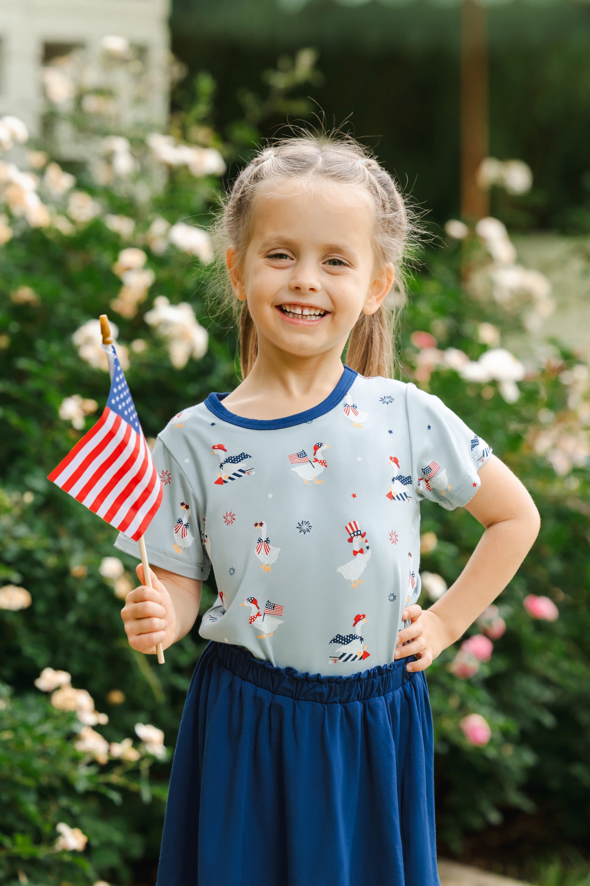 Child wearing light blue Toddler Basic Tee in Patriotic Goose print over a Tahoe skort holding a flag outdoors