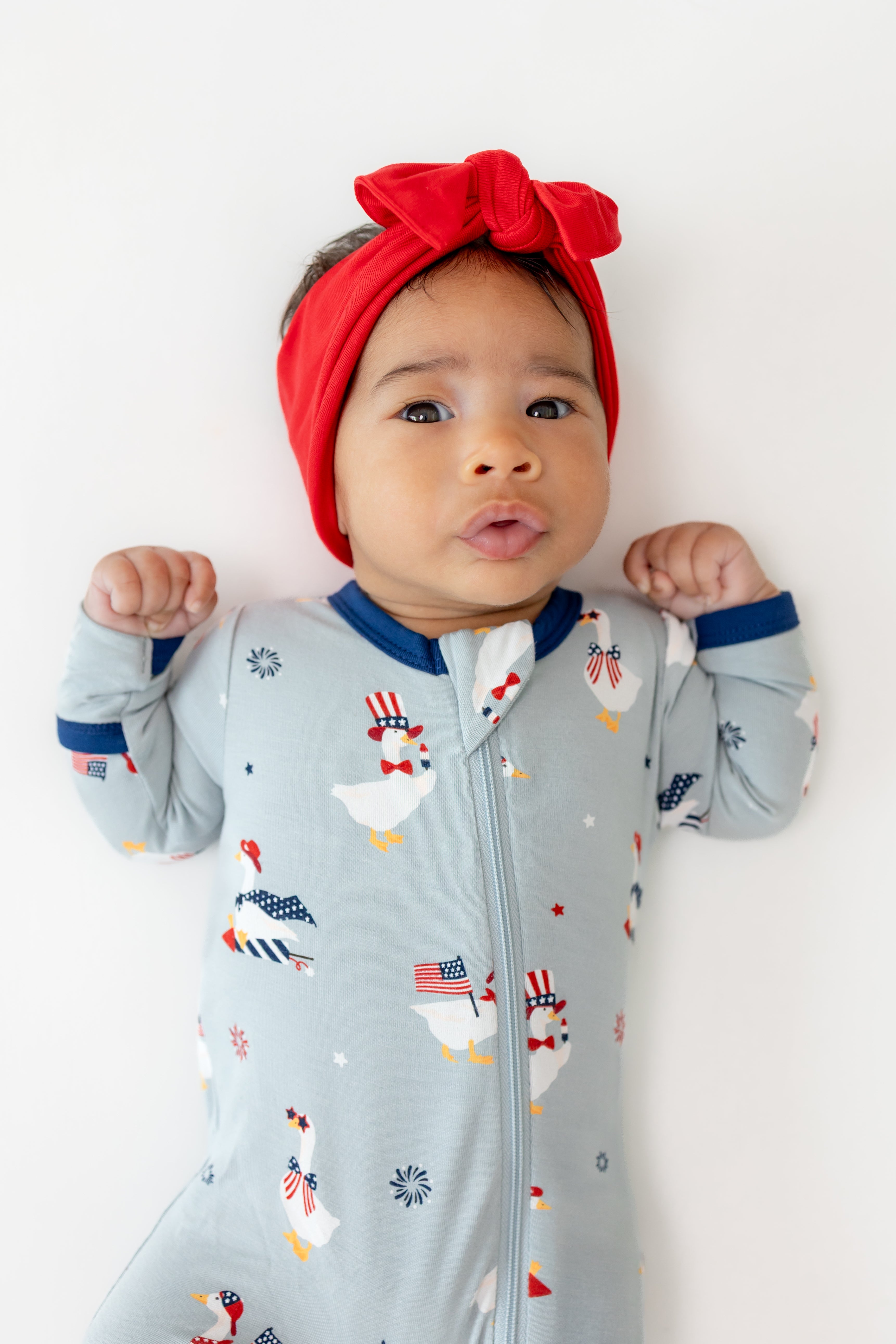 Baby wearing Patriotic Goose print Light blue Zippered footie with a red bow headband on a white background