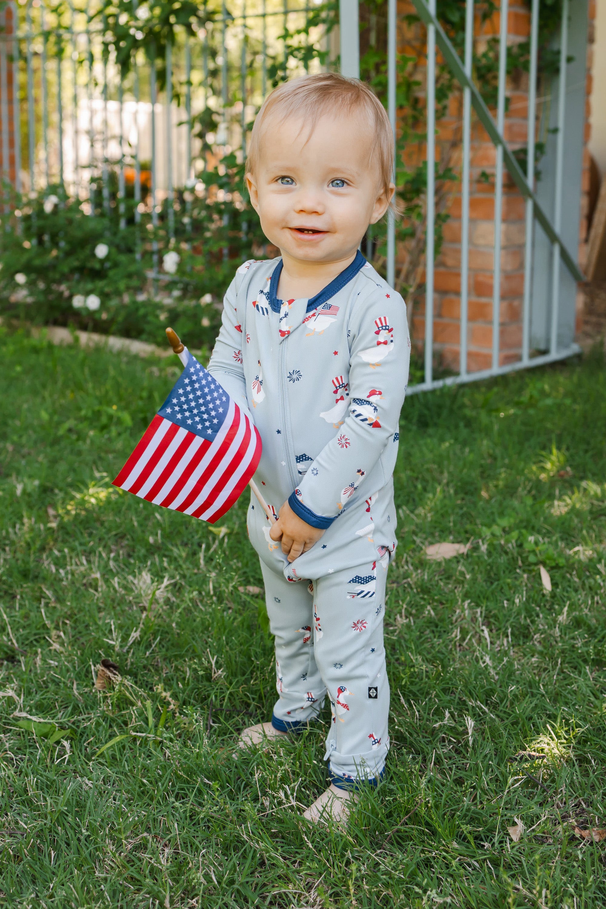 Child standing wearing a light blue zippered romper in Patriotic Goose print with a flag outdoors