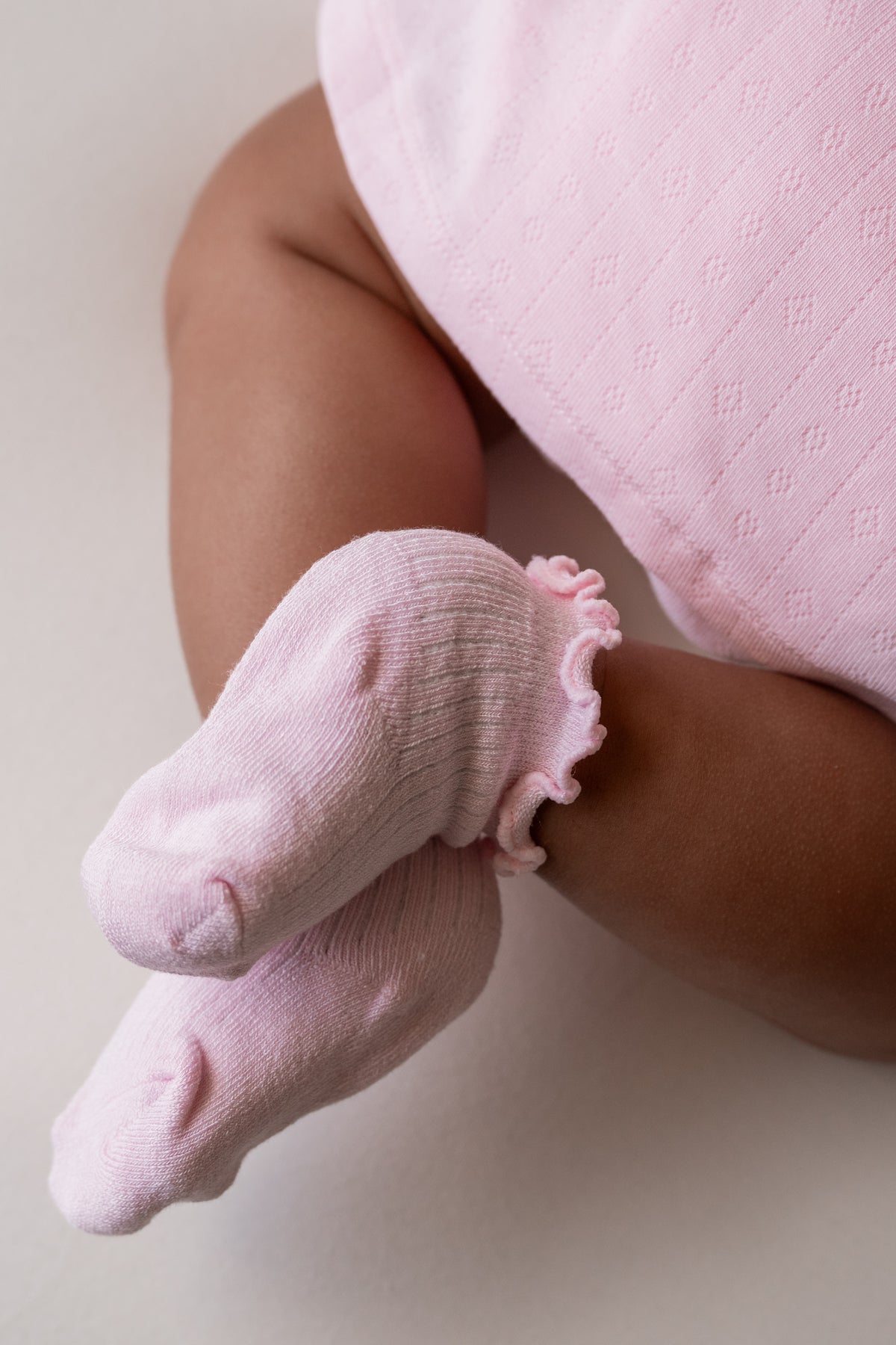 A close-up of a baby's legs and feet wearing light pink ribbed socks with ruffled trim, paired with a matching pink outfit against a soft neutral background.
