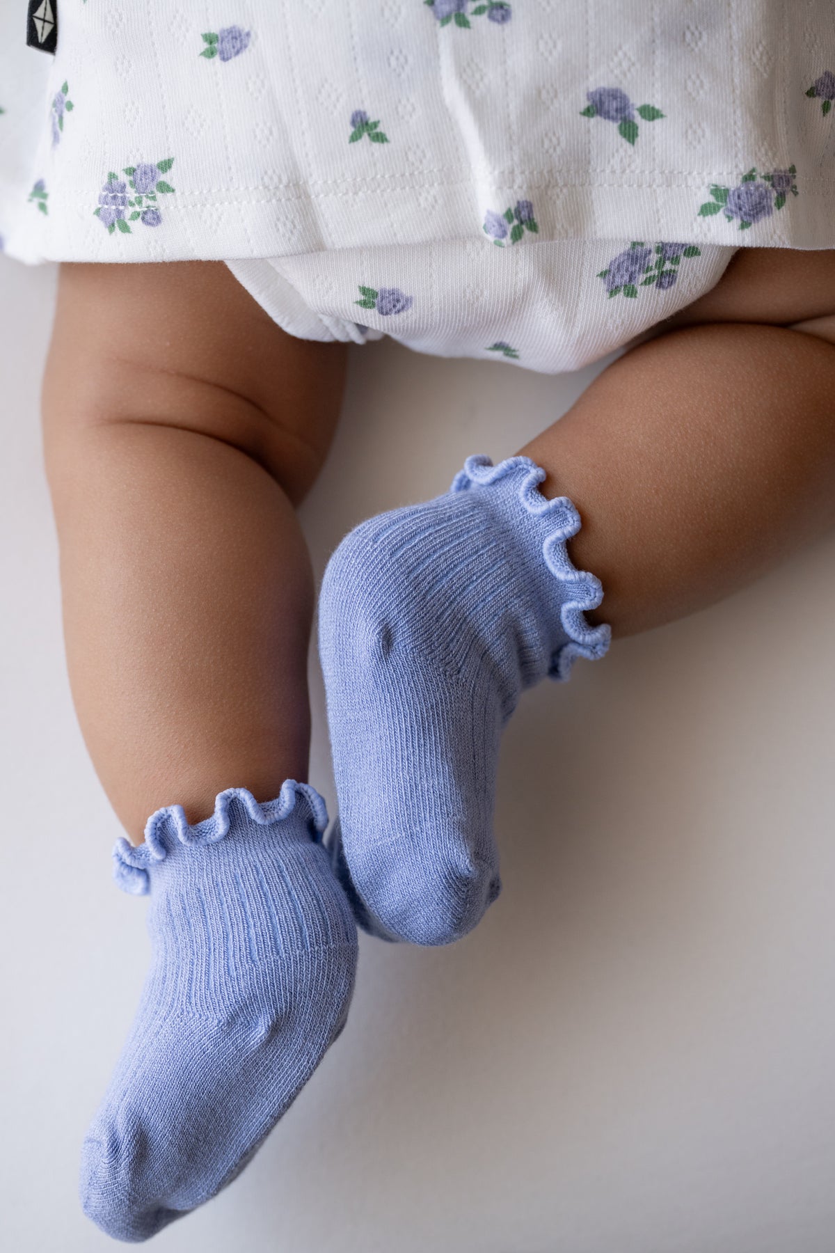 A close-up of a baby's legs wearing light blue ruffled socks, with the baby's white floral dress visible at the top against a plain light background.