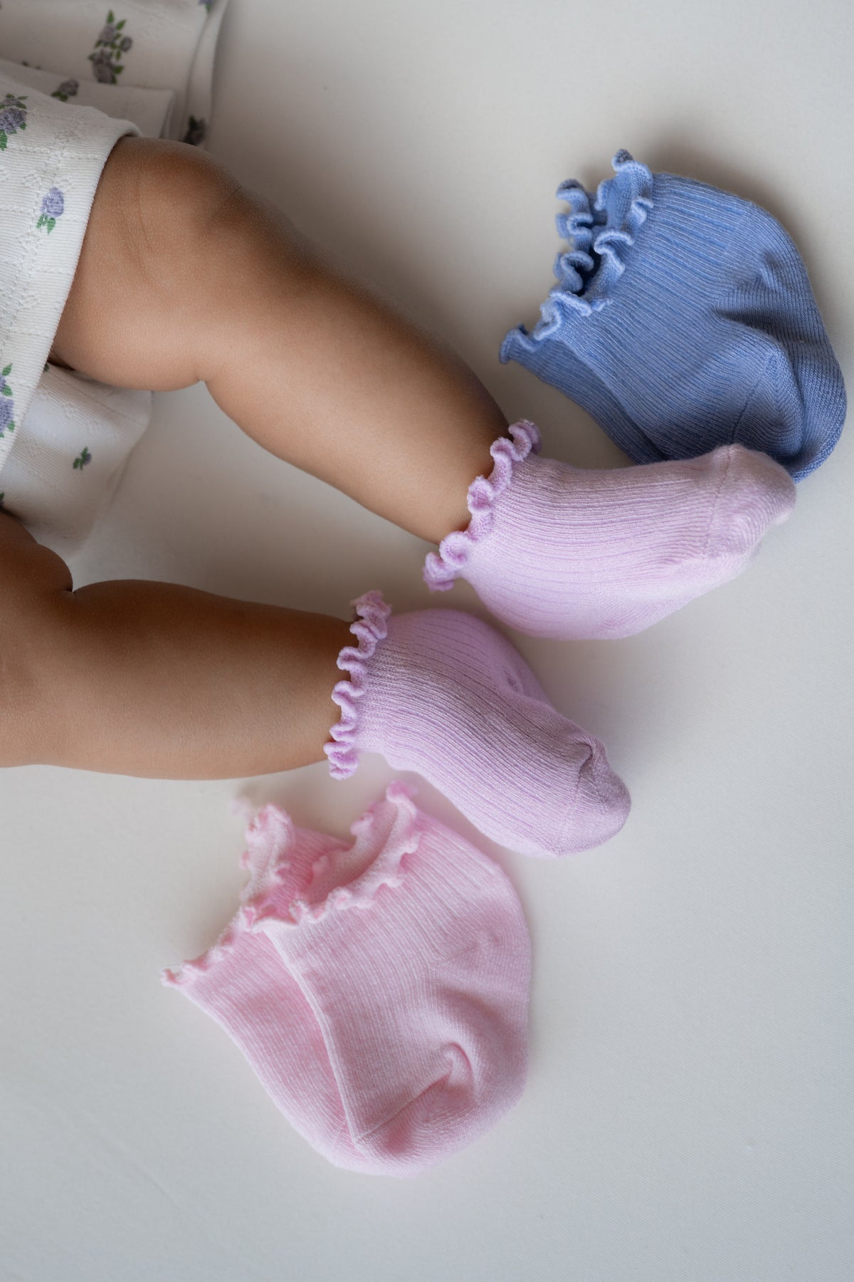 Baby wearing light purple ruffle socks with 2 other socks laying next to them in a periwinkle and light pink color