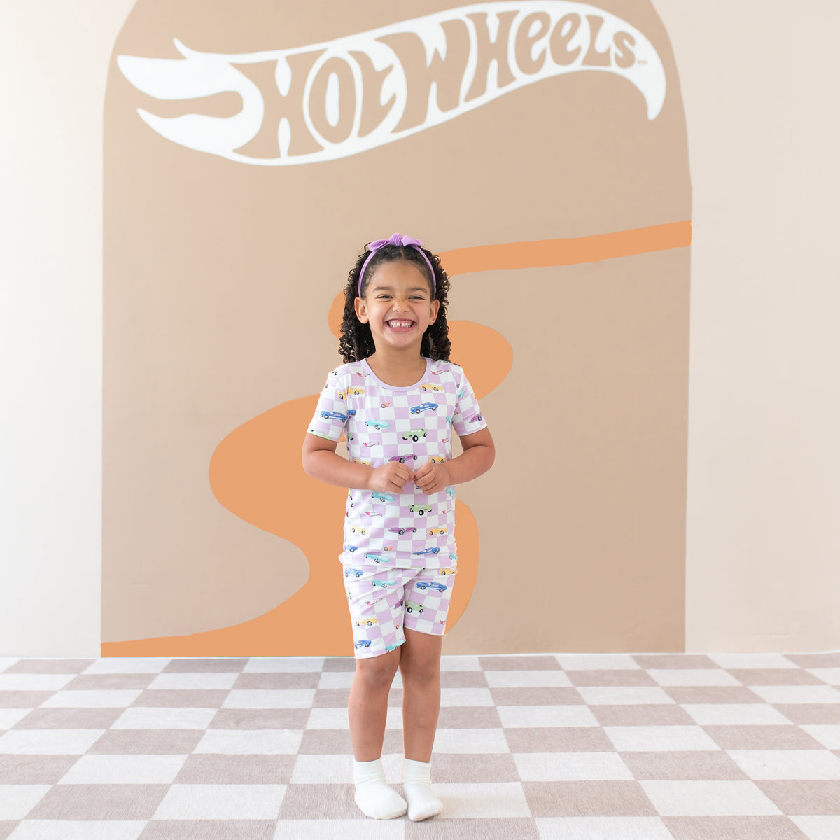 Smiling girl wearing the Short Sleeve Pajamas in Sparkles and Speed standing in front of Hot Wheels™ back drop