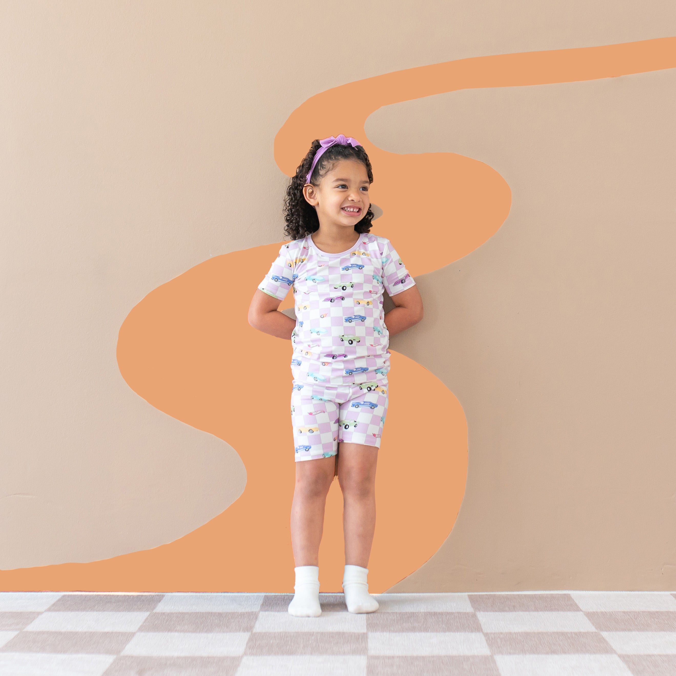 Smiling girl looking off the side with her hands behind her back standing in front of a back drop wearing the Short Sleeve Pajamas in Sparkles and Speed