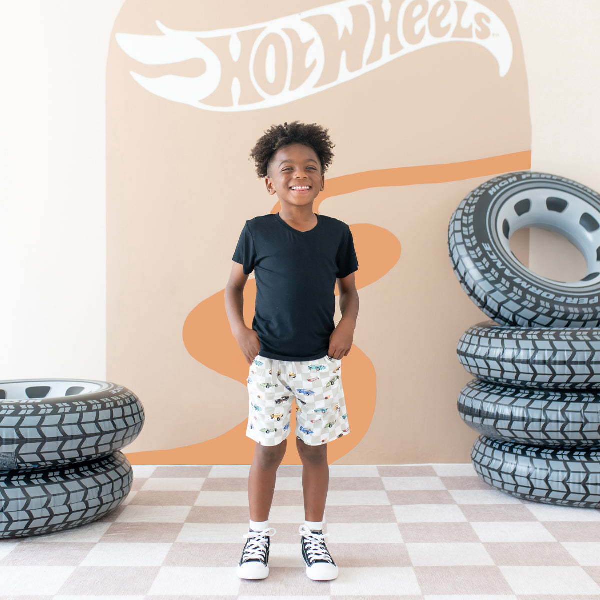 Young Smiling boy wearing the Drawstring Short in Fast and Fierce paired with a Toddler V-Neck in Midnight standing in front of a back drop with inflatable tires