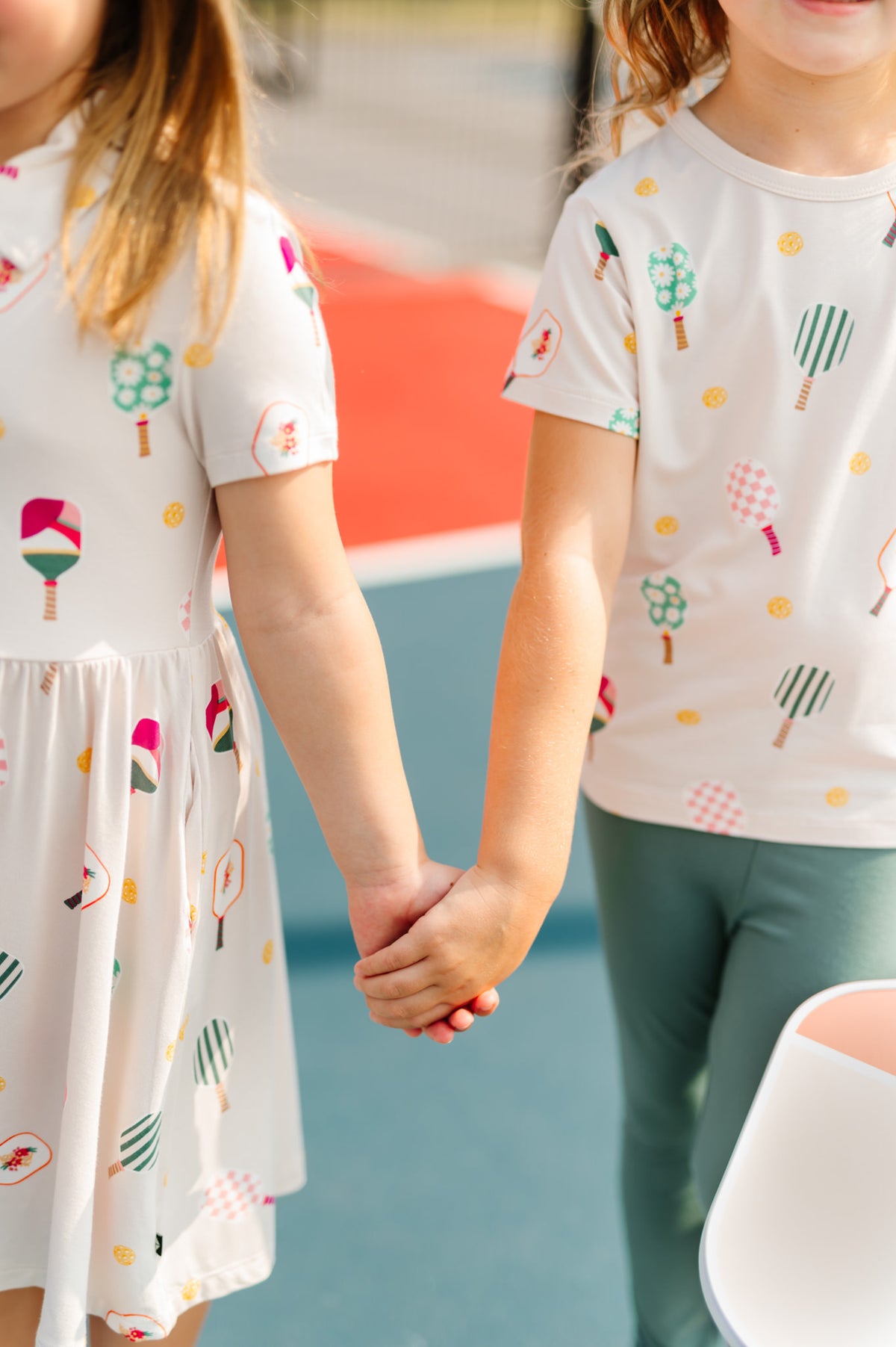 Close up of two girls on pickleball court holding hands wearing Polo Dress and Toddler Basic Tee in Pickleball print