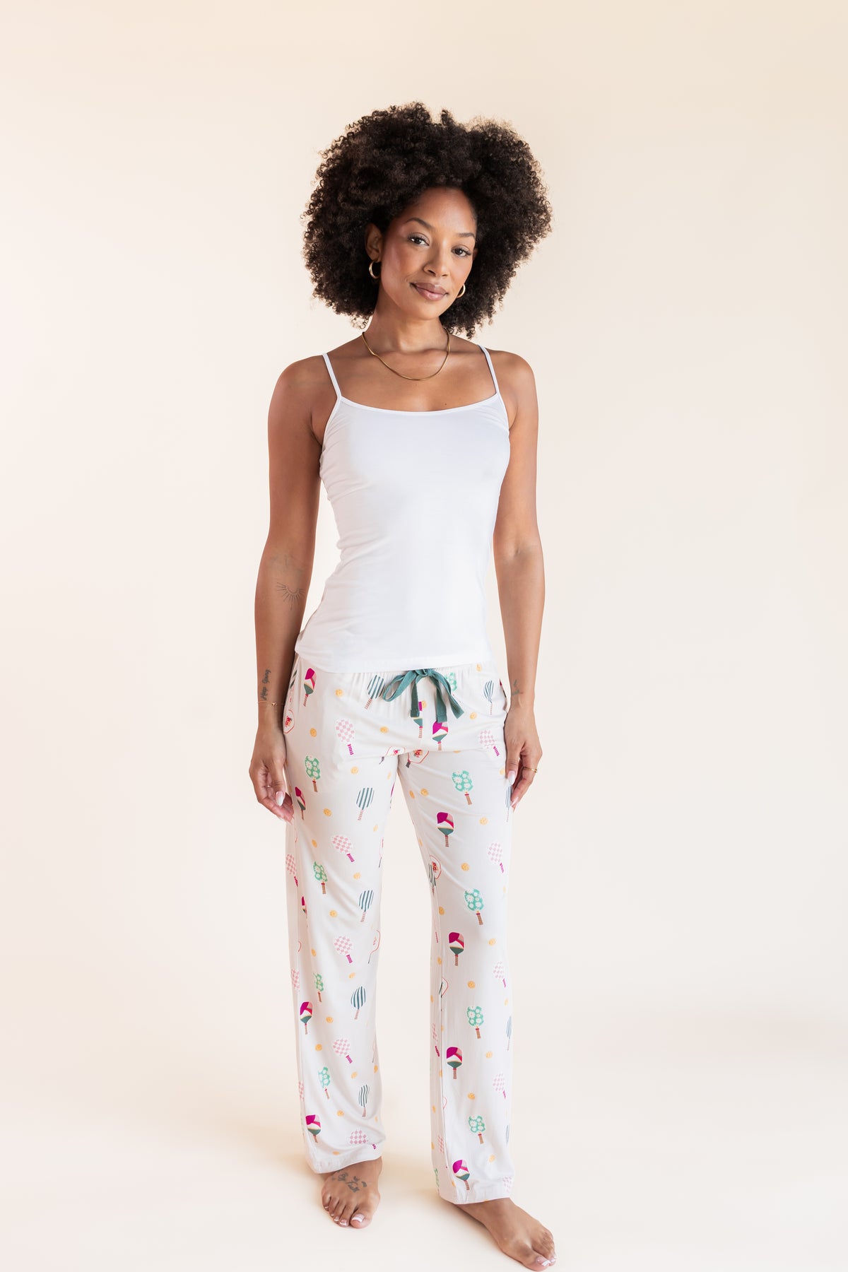 A woman modeling a two-piece pajama set consisting of a white camisole top and white pants with a colorful pickleball print, set against a solid cream background.