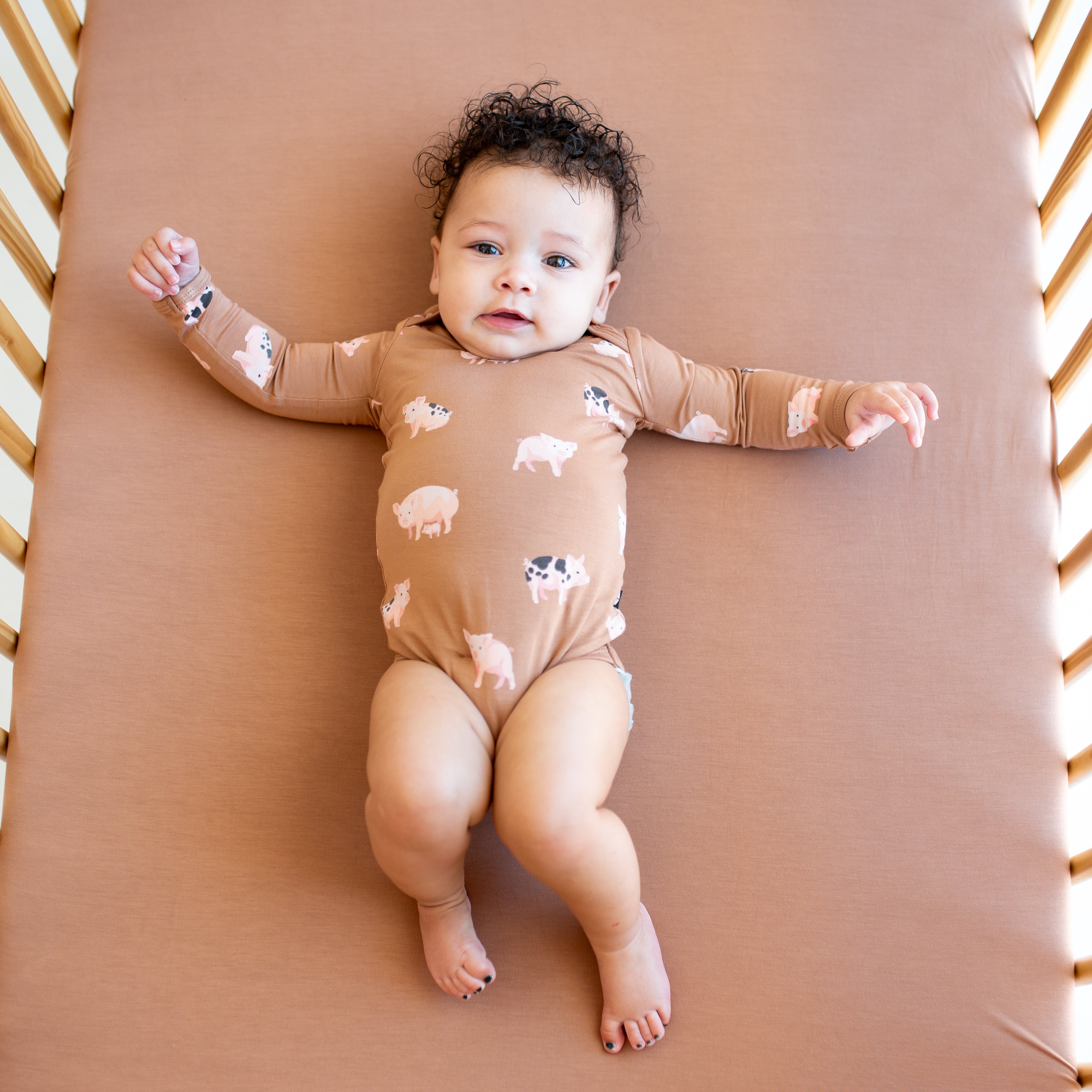 Infant laying in a crib on a Latte crib sheet wearing the Long Sleeve Bodysuit in Pig
