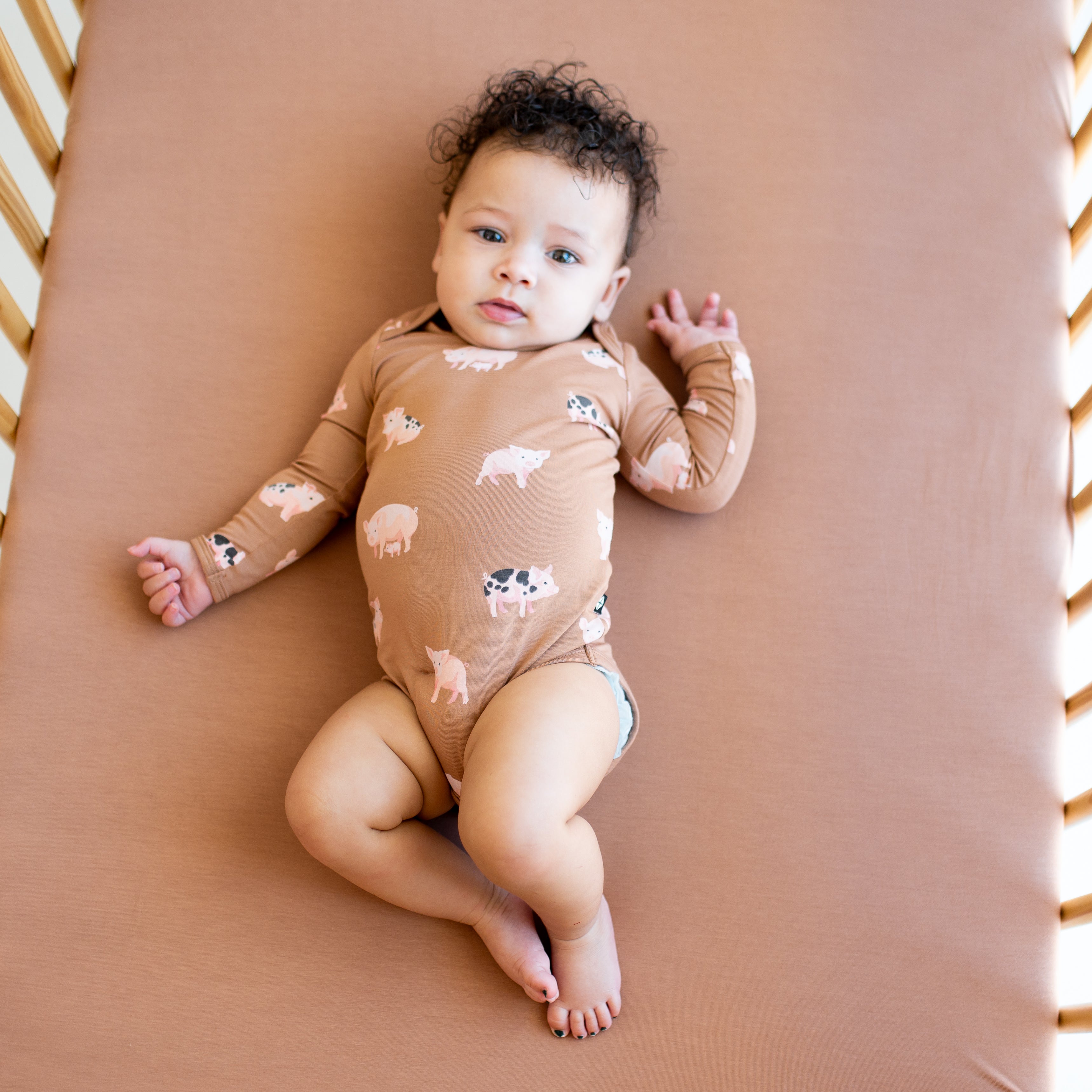 Infant laying in a crib on a latte crib sheet wearing the Long Sleeve Bodysuit in Pig
