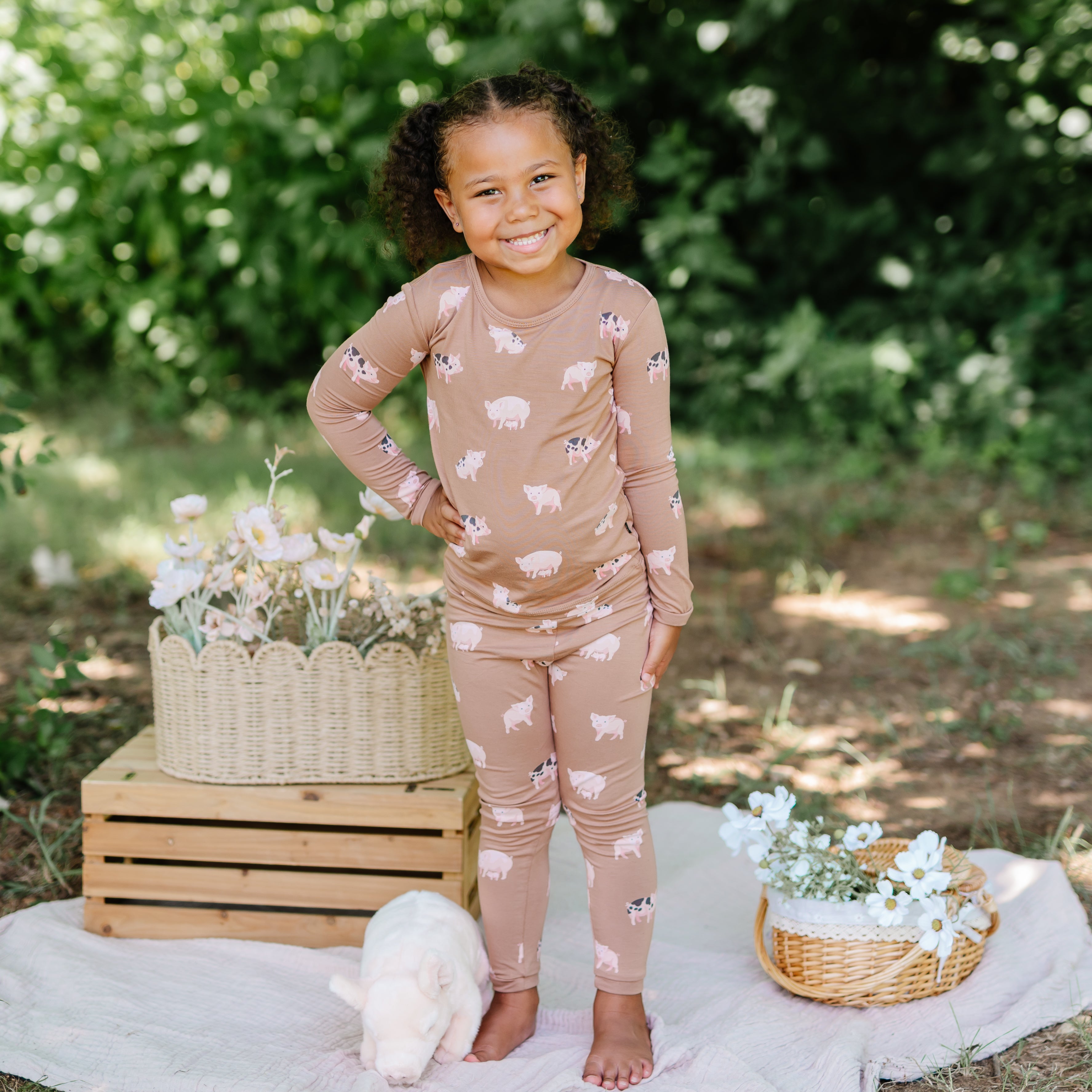 Young girl standing outside with one hand on hip between flowers in wicker baskets, beside a stuffed toy pig on a white blanket wearing the Long Sleeve Pajamas in Pig