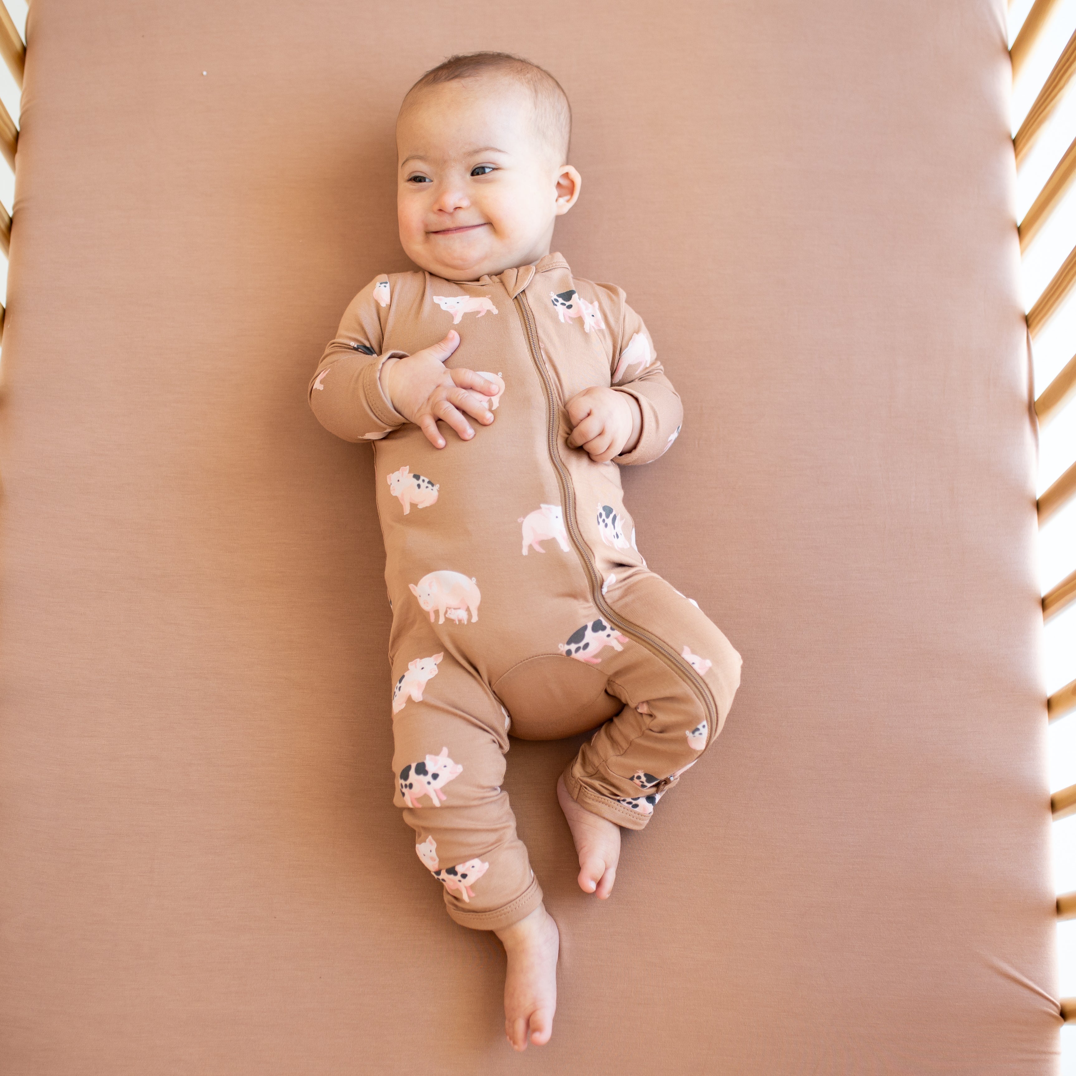 Smiling infant laying in a crib on top of a latte crib sheet wearing the Zippered Romper in Pig