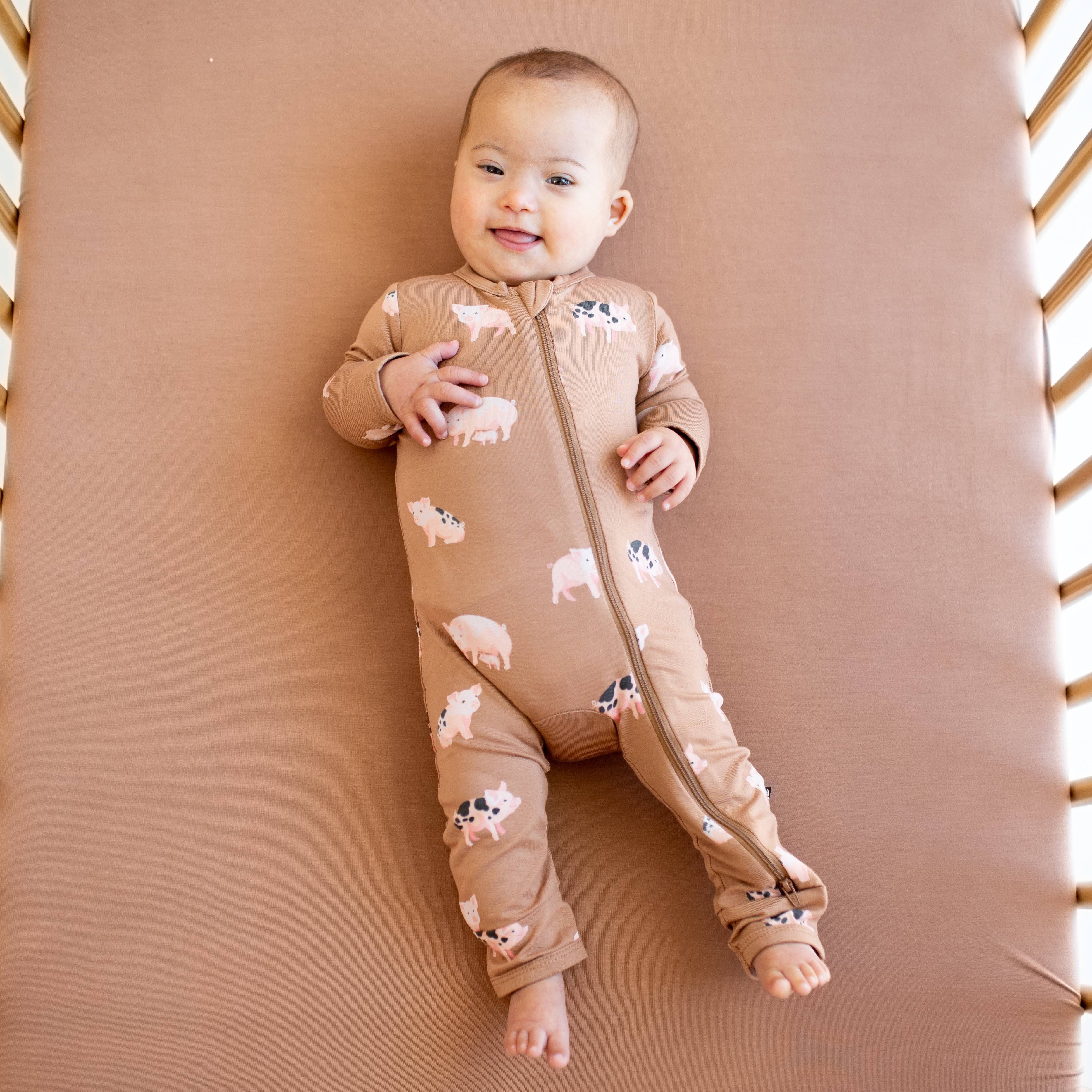 Infant sticking her tongue out laying in a crib on top of a latte crib sheet wearing the Zippered Romper in Pig
