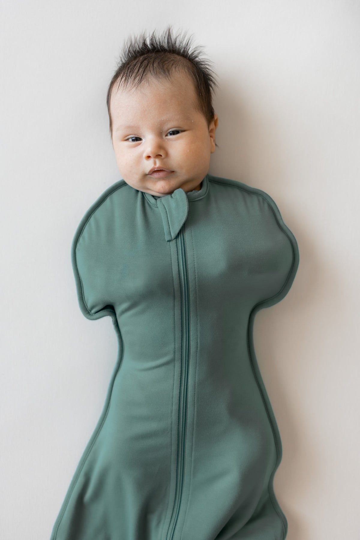 A baby wearing a sage green zip-up baby swaddle bag, lying on their back against a plain off-white wall.