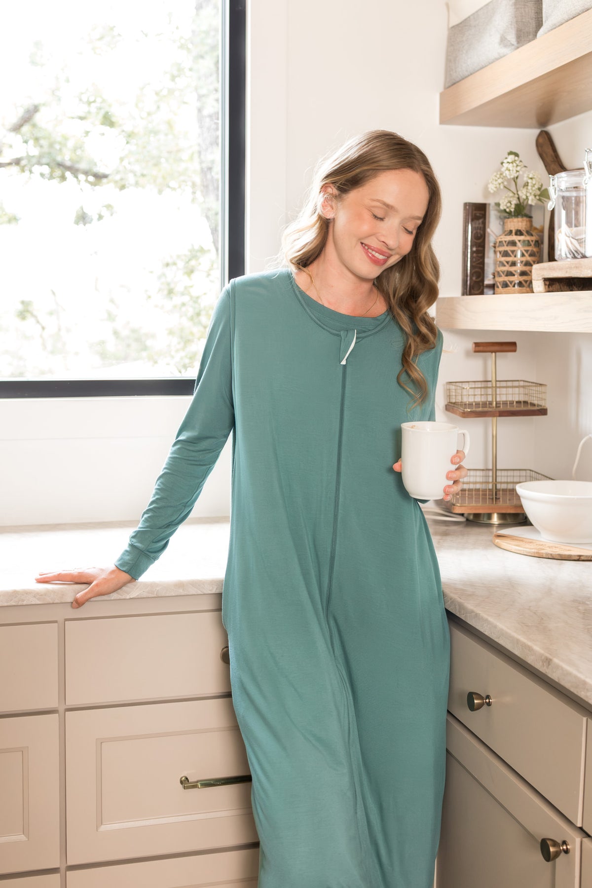 A woman wearing a sage green sleep sack in a bright kitchen, holding a white mug.