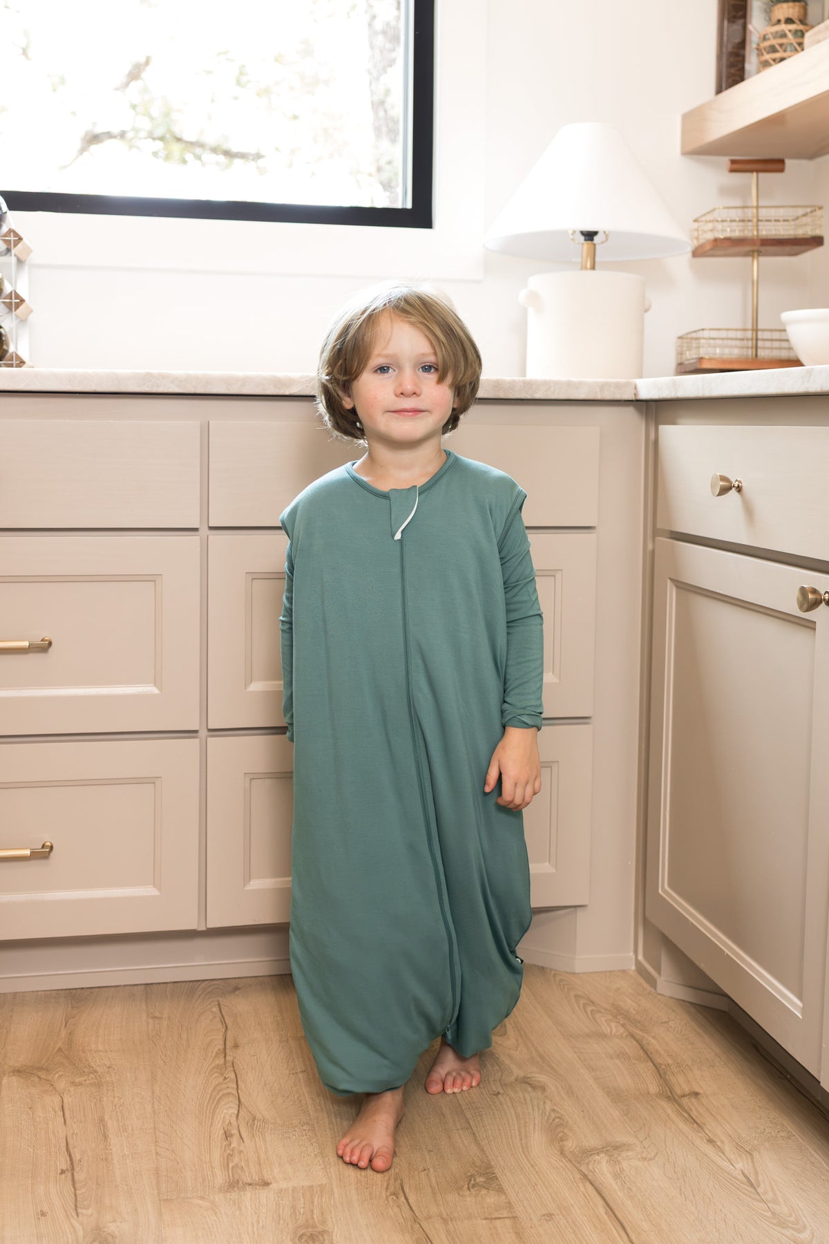 A young child wearing a sage green footless sleep sack stands barefoot in a bright kitchen with cream-colored cabinets and light wood floors.