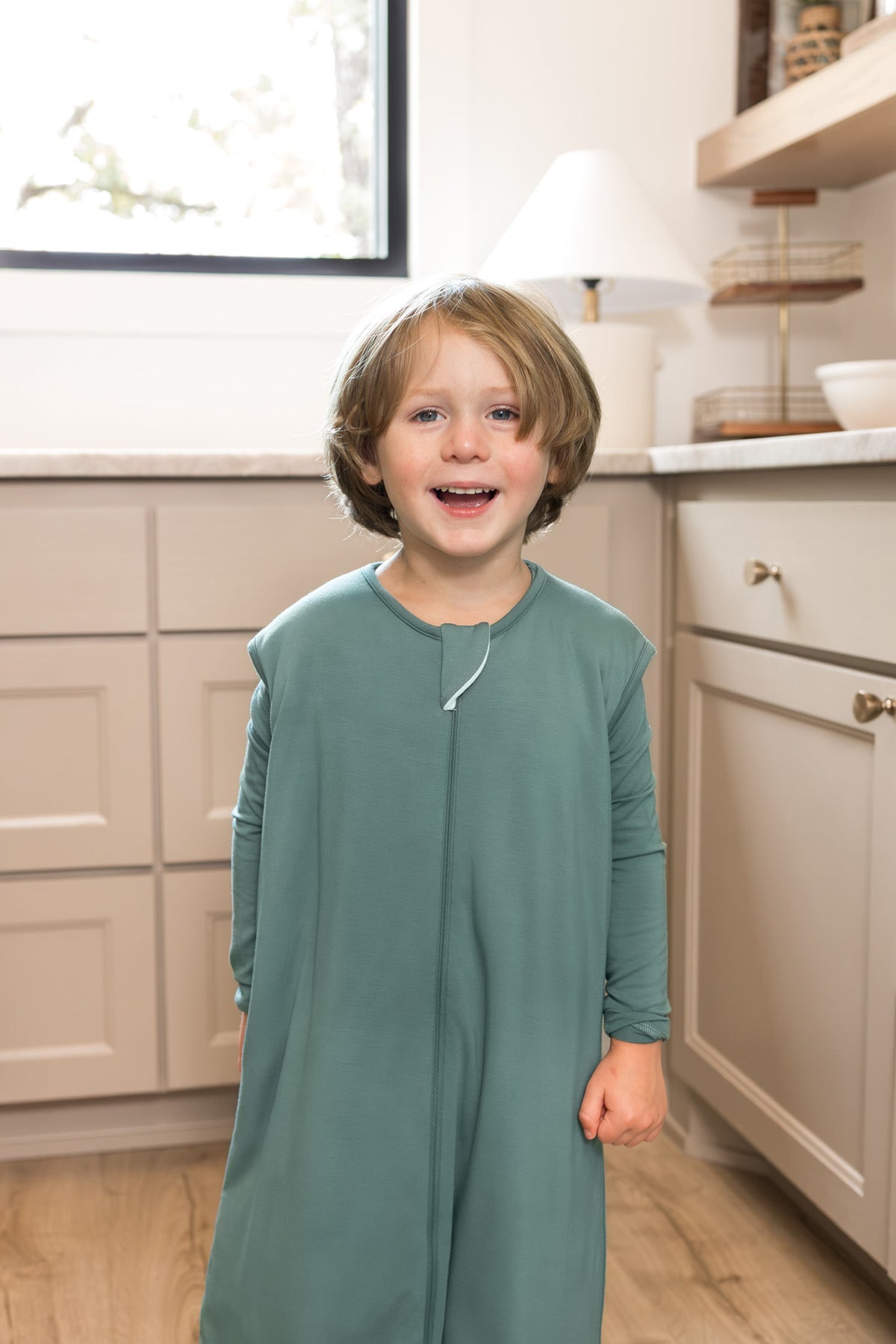 A young child in a kitchen wearing a sage green sleep bag walker. The background features cream-colored cabinets, a light wood floor, and a window.