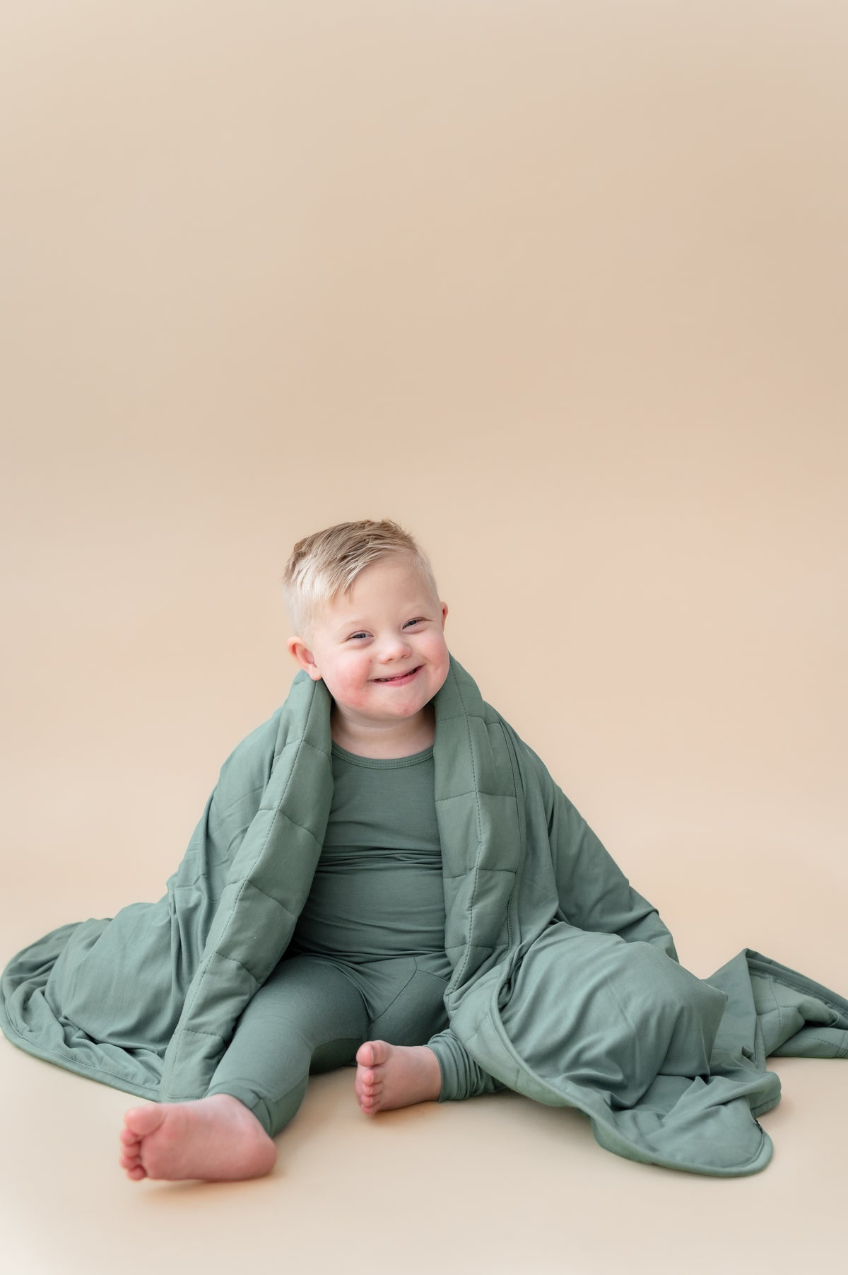 Toddler Blanket in Pine 2.5 wrapped over toddler's shoulder wearing similar color long sleeve pajama set on a beige background