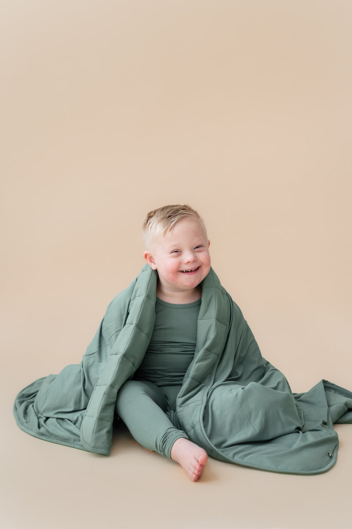 Toddler sitting with Toddler Blanket in Pine 2.5 wrapped over his shoulder wearing similar color long sleeve pajama set on a beige background