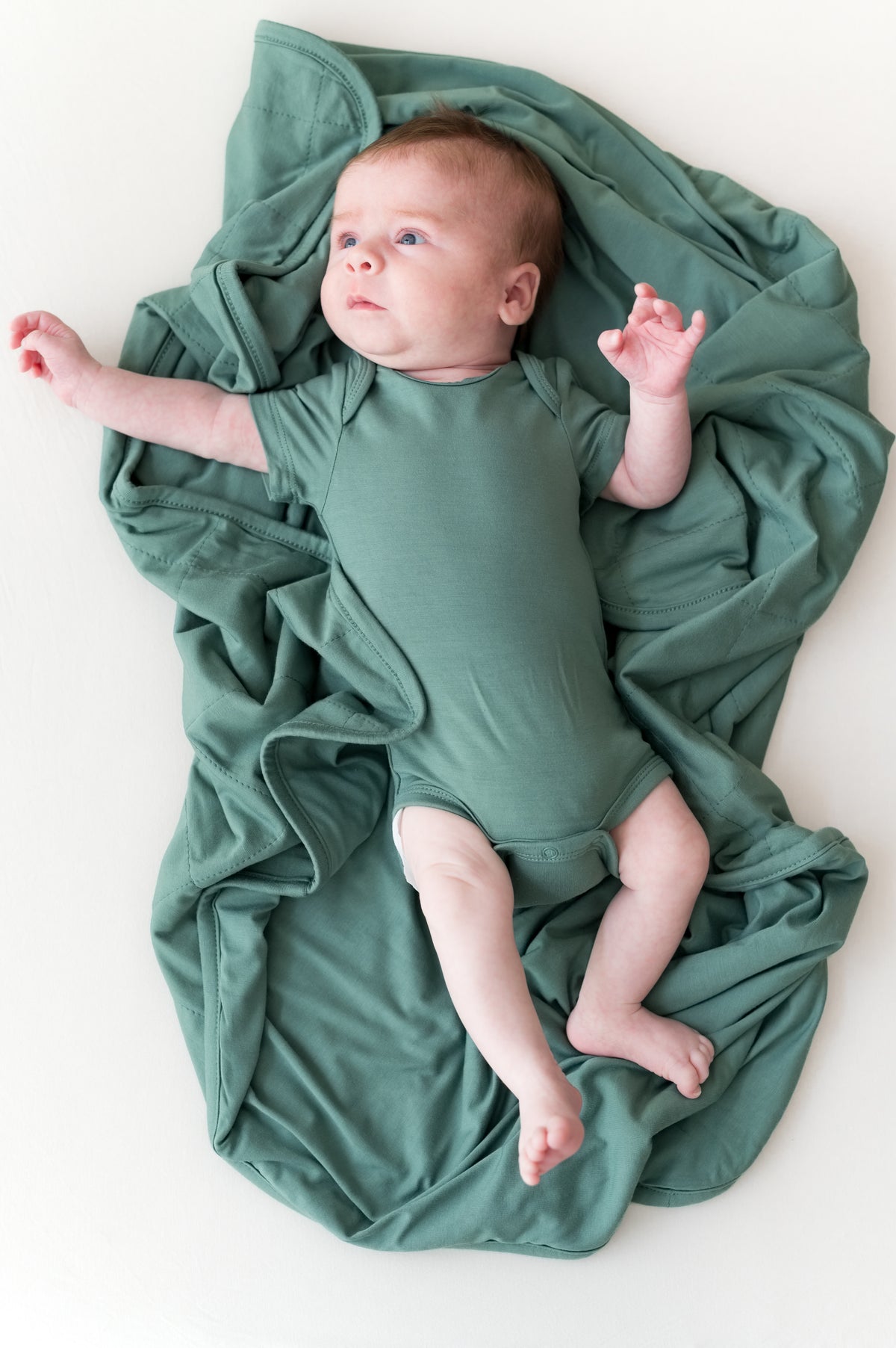 Baby lying on Baby Blanket in Pine wearing a similar color bodysuit on a beige background