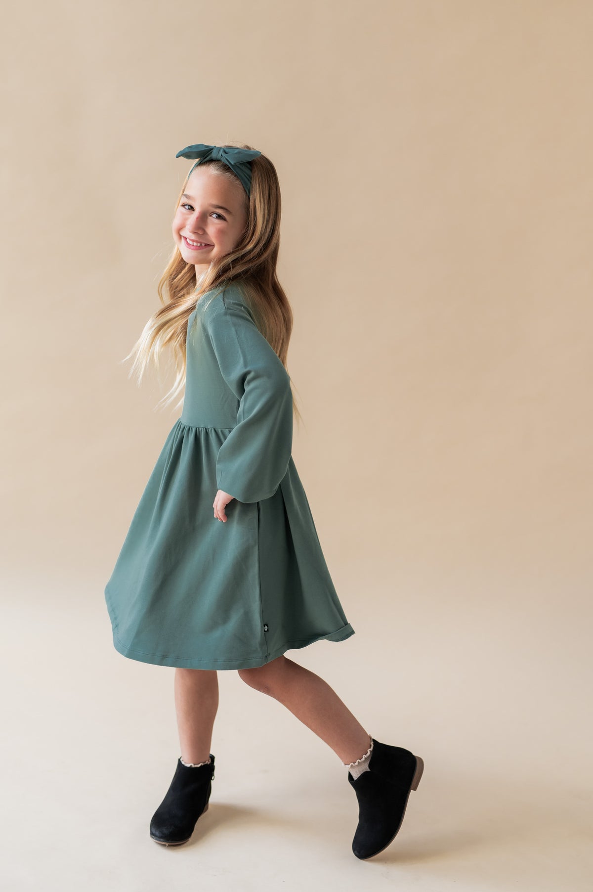 Side pose of Girl wearing Long Sleeve Skater Dress in Pine color with similar color knotted bow headband on a beige background