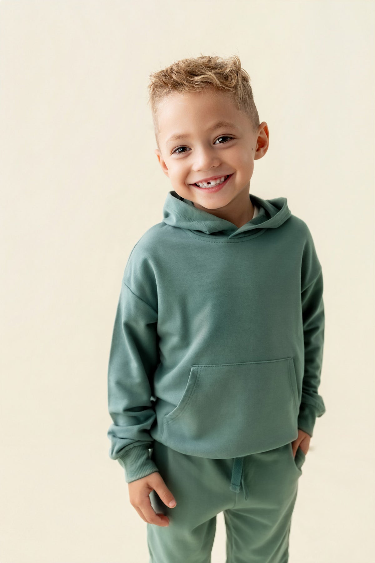 Boy smiling in Hoodie in Pine color and pine pants on a beige background