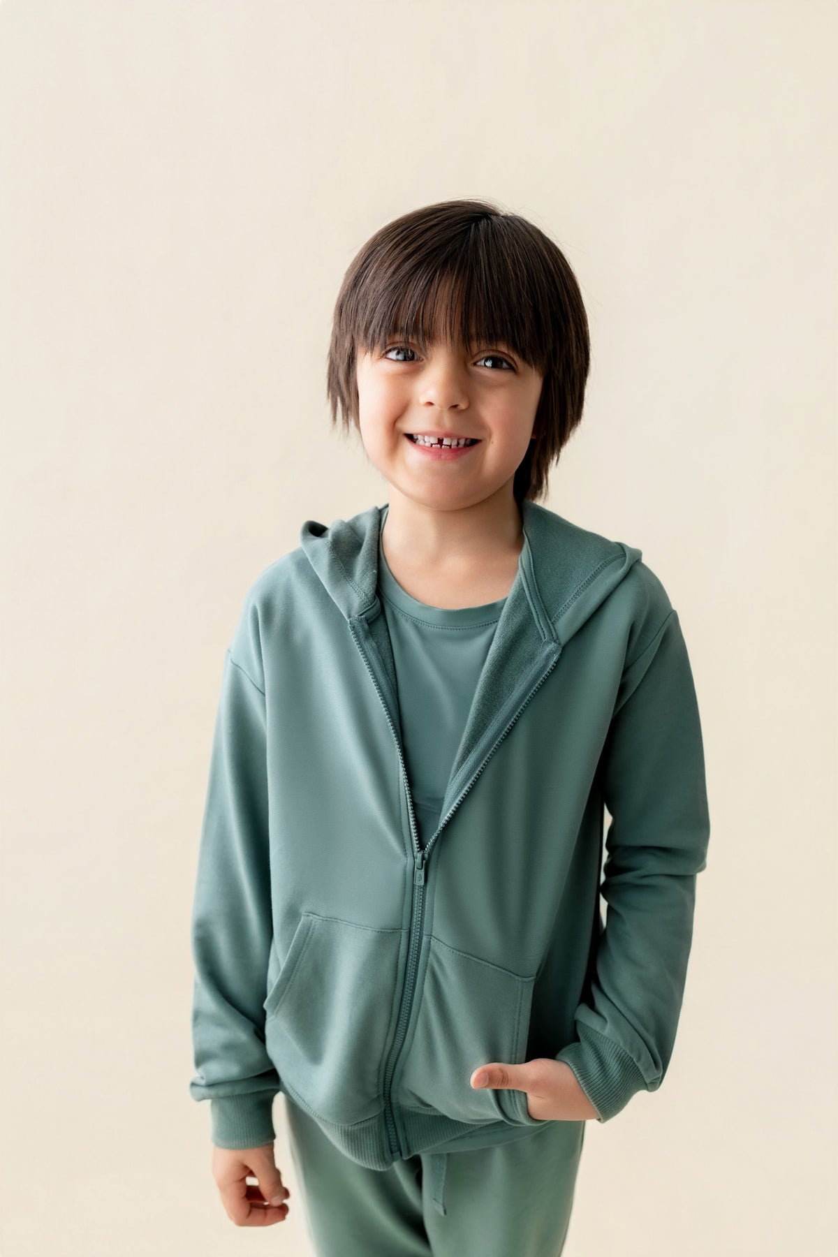 Boy posing in Zip-Up Hoodie in Pine color with pockets over similar color jogger pant on a beige background