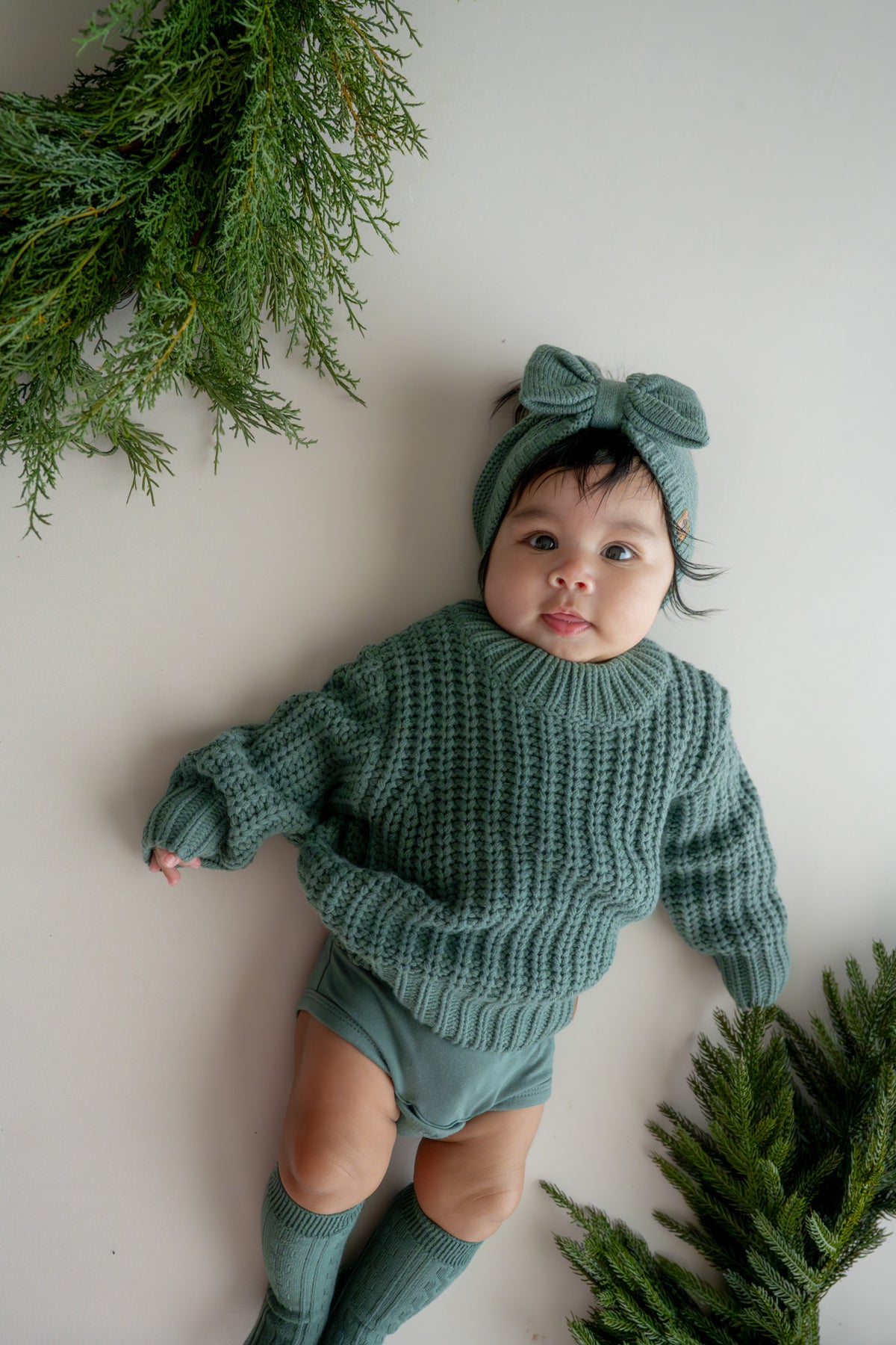 Baby wearing Chunky Knit Sweater, chunky knit bow headband in Pine and socks in pine lying on a beige background