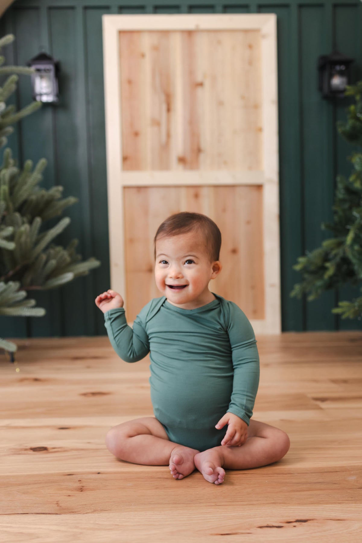 A smiling baby sitting cross-legged on a wooden floor, wearing a long-sleeve sage green bodysuit. The background features a rustic wooden door set against a dark green paneled wall, with decorative lights and greenery on either side.