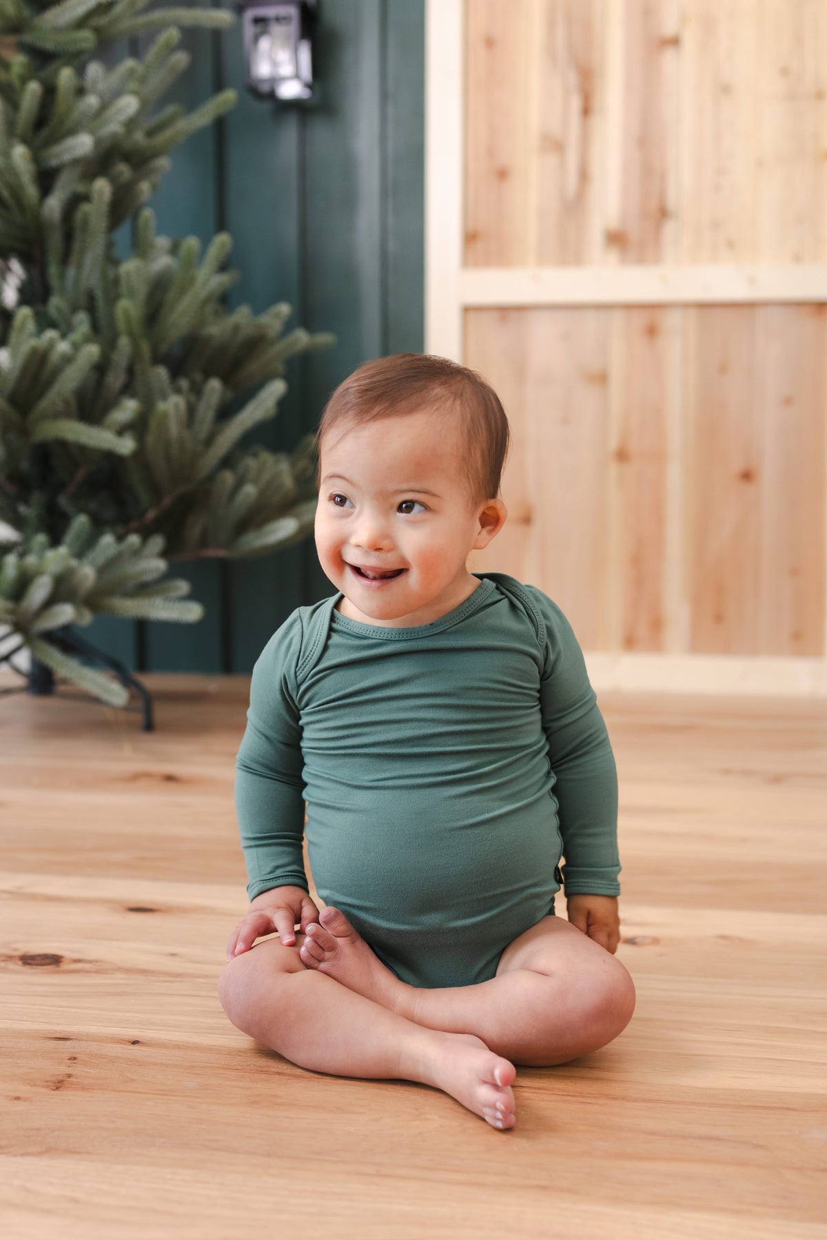 A child wearing Long Sleeve Bodysuit in Pine sitting on wooden floor
