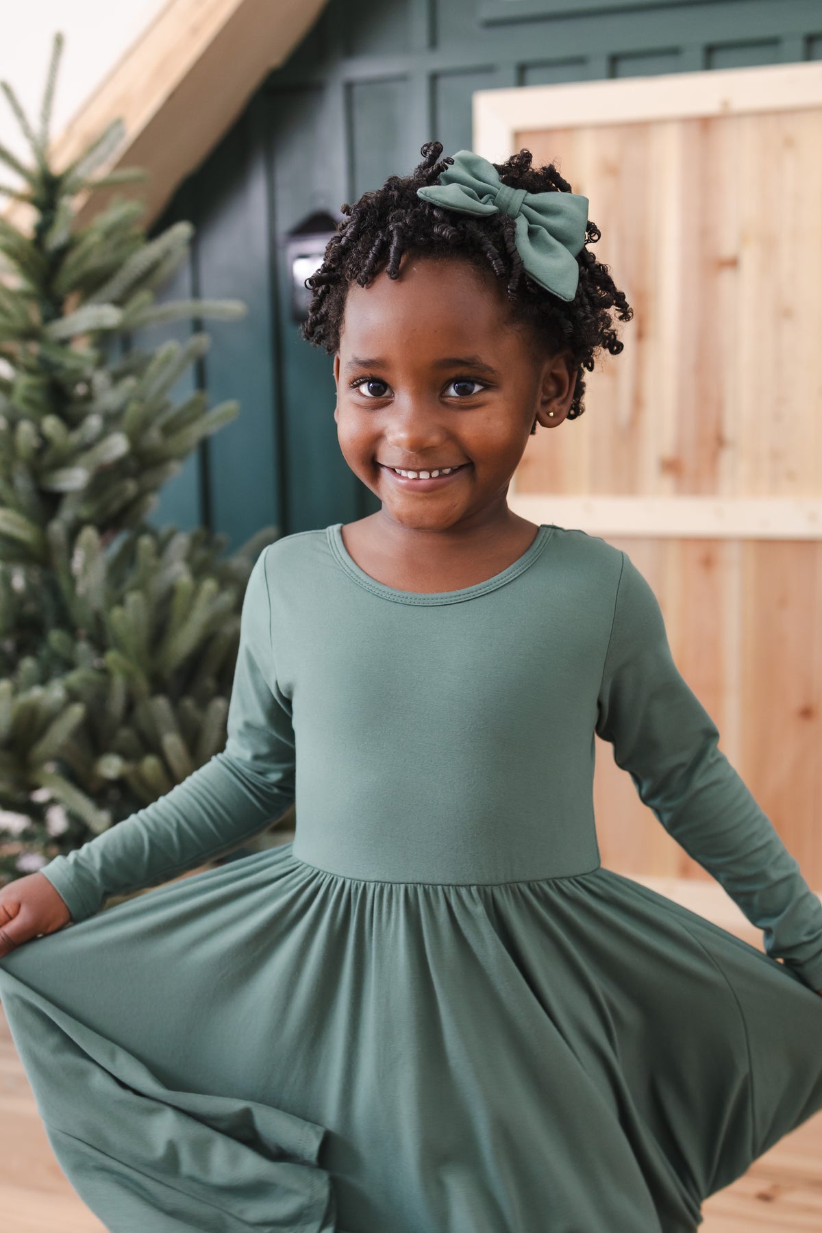 Girl showcasing Long Sleeve Twirl Dress in Pine standing in front of a beige door