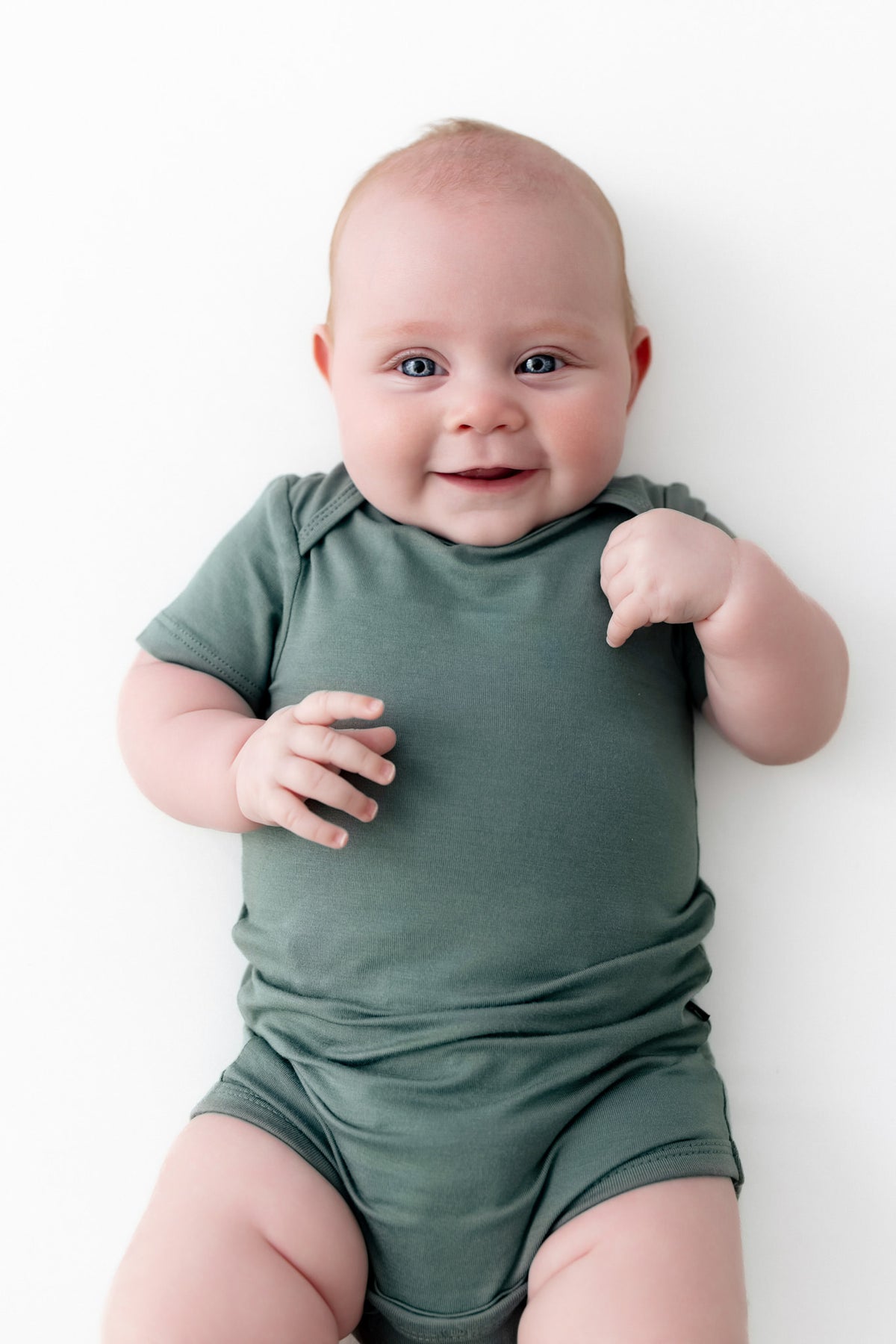 A smiling baby lying on their back wearing a sage green short-sleeve bodysuit against a clean white background.