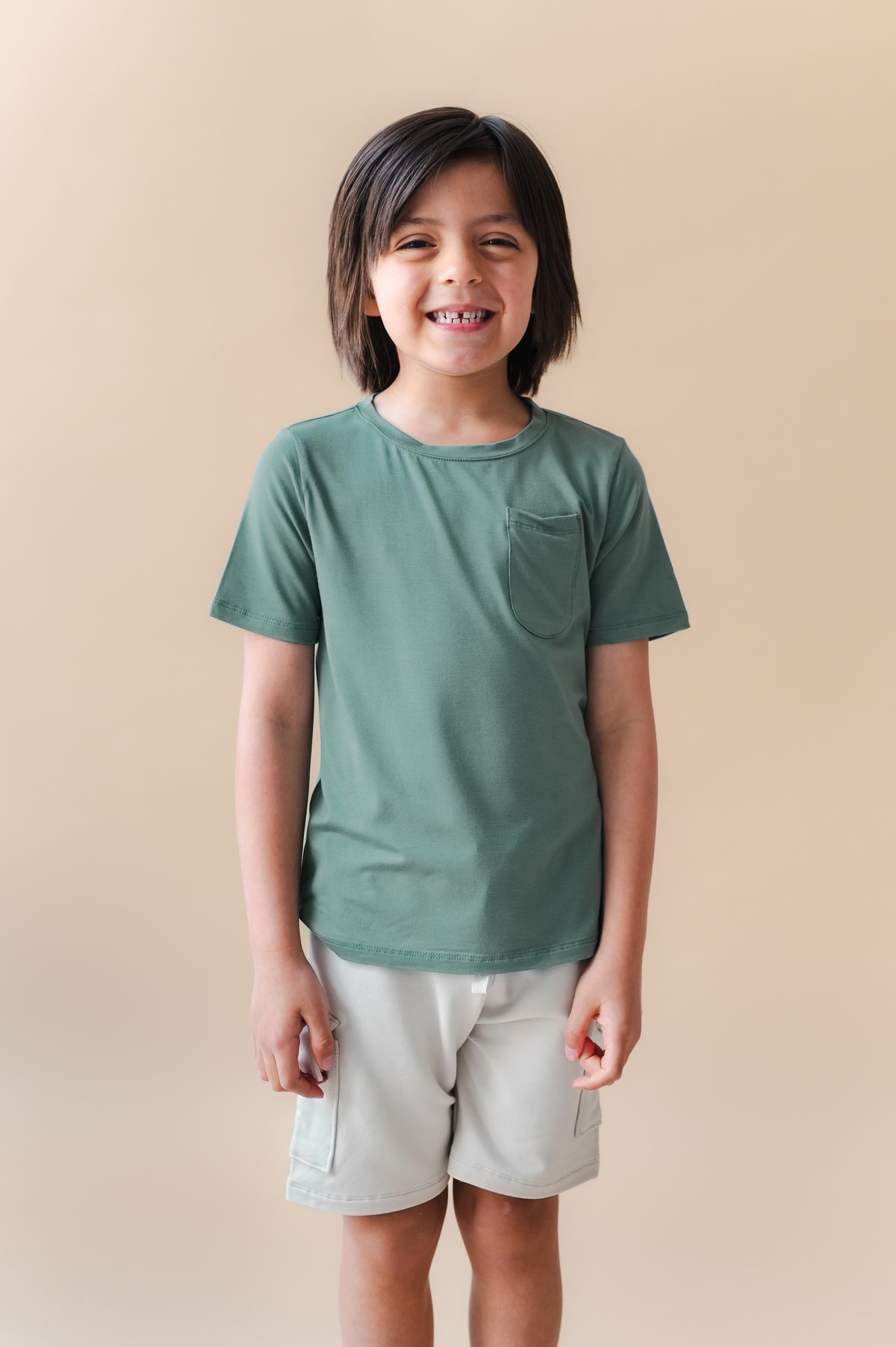 Boy wearing Toddler Crew Neck Tee in Pine over white shorts on a beige background